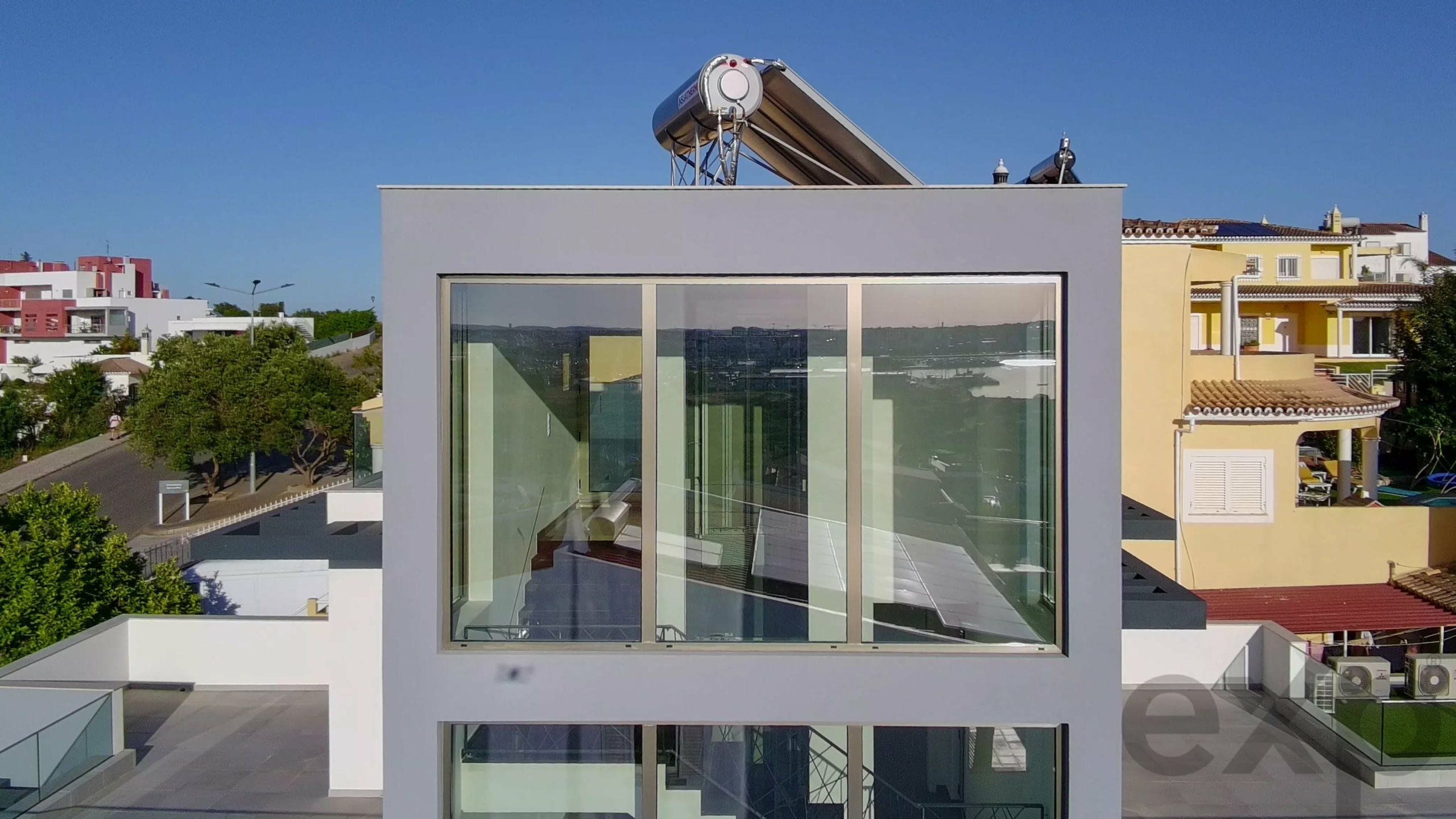 Modern rooftop with large glass facade and a solar water heater on top, overlooking a sunny residential neighborhood.