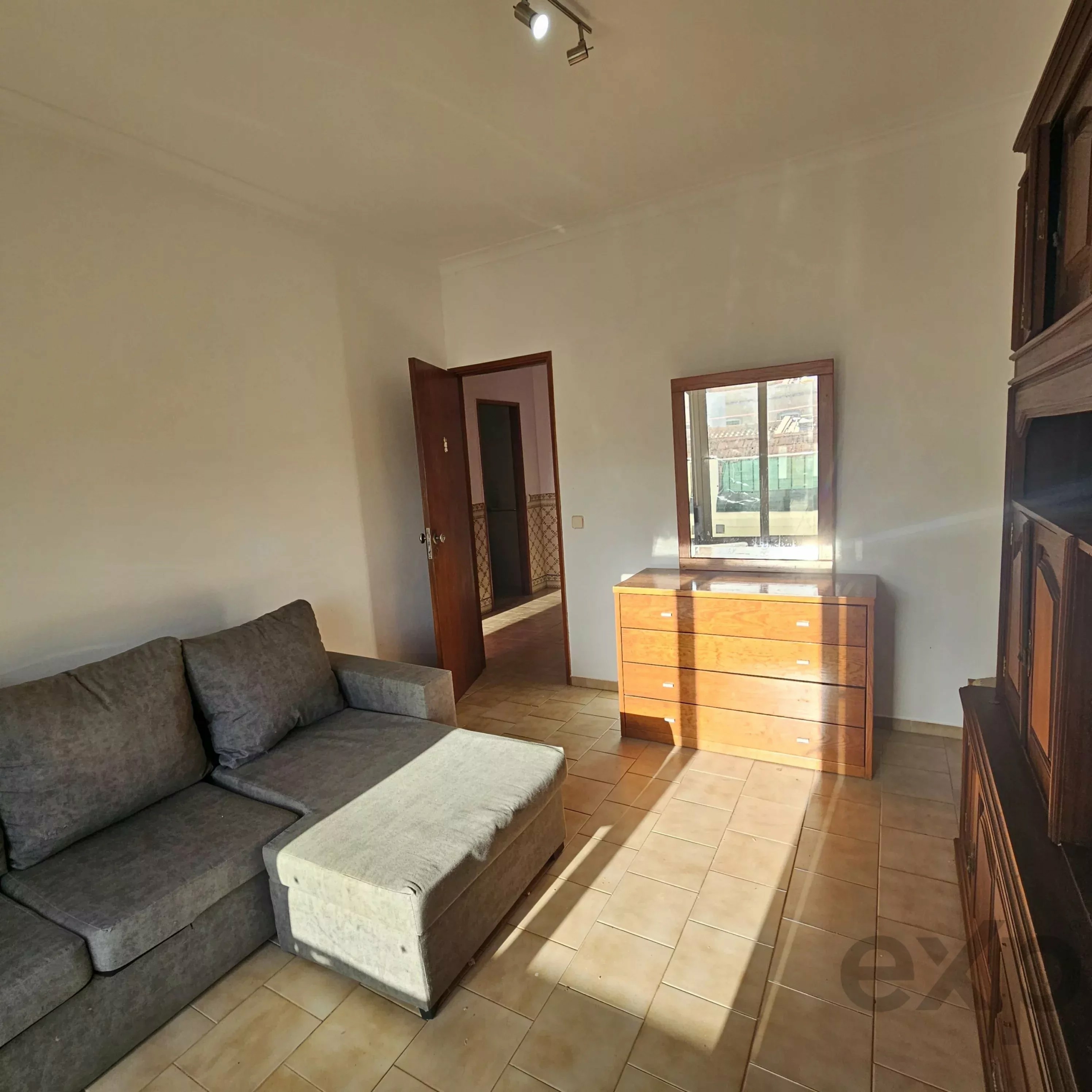 Living room with gray sectional sofa, wooden dresser, and large windows letting in natural light; tiled floor and doorway visible on the left.