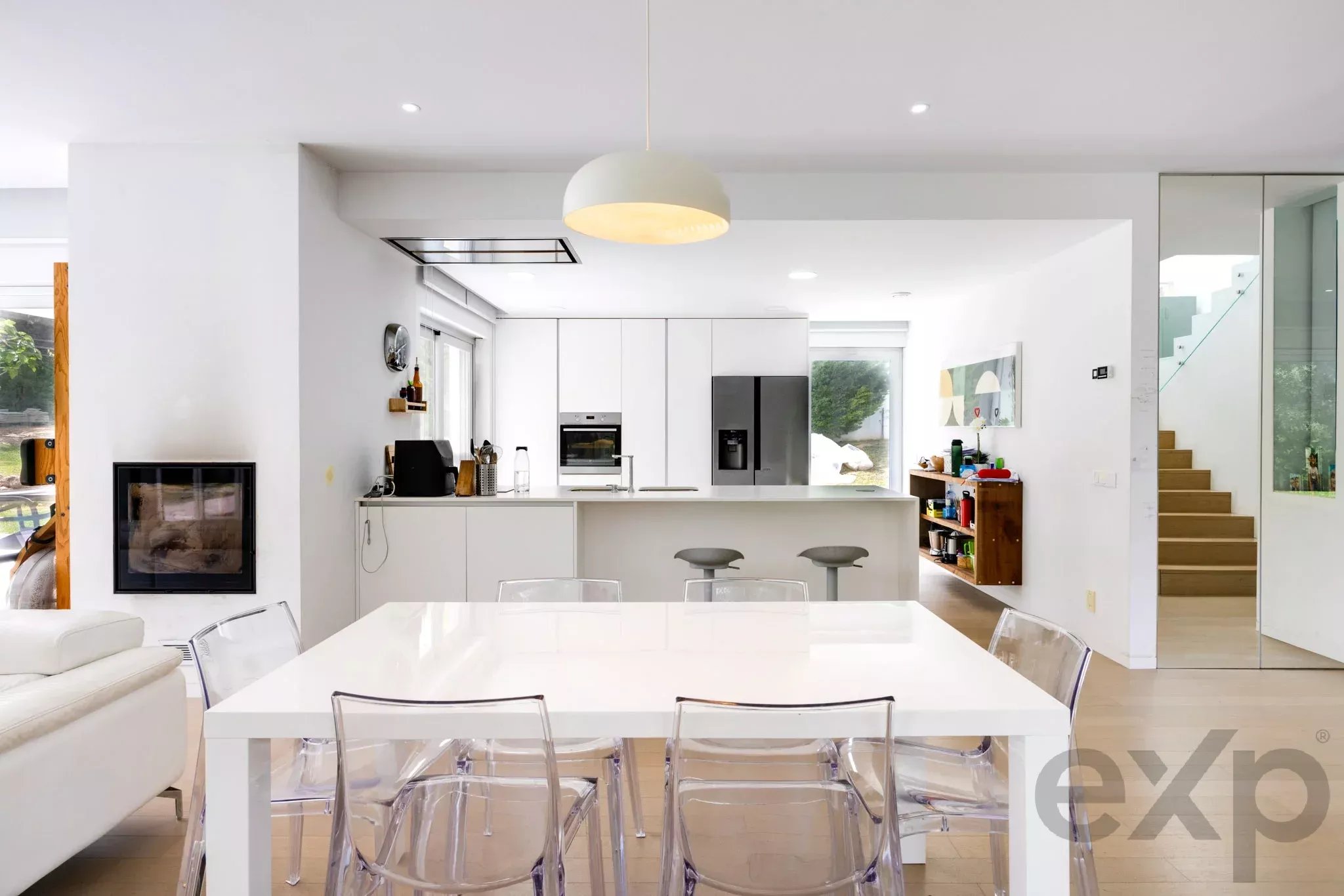 Bright, minimalist kitchen and dining area with white cabinets, a long white island, and clear acrylic chairs around a large dining table under a pendant light.
