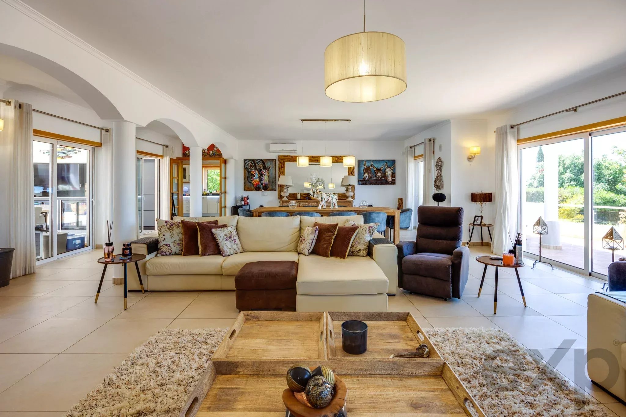 Bright open-plan living room with a beige sofa, patterned cushions, and a large wooden coffee table under pendant lights.