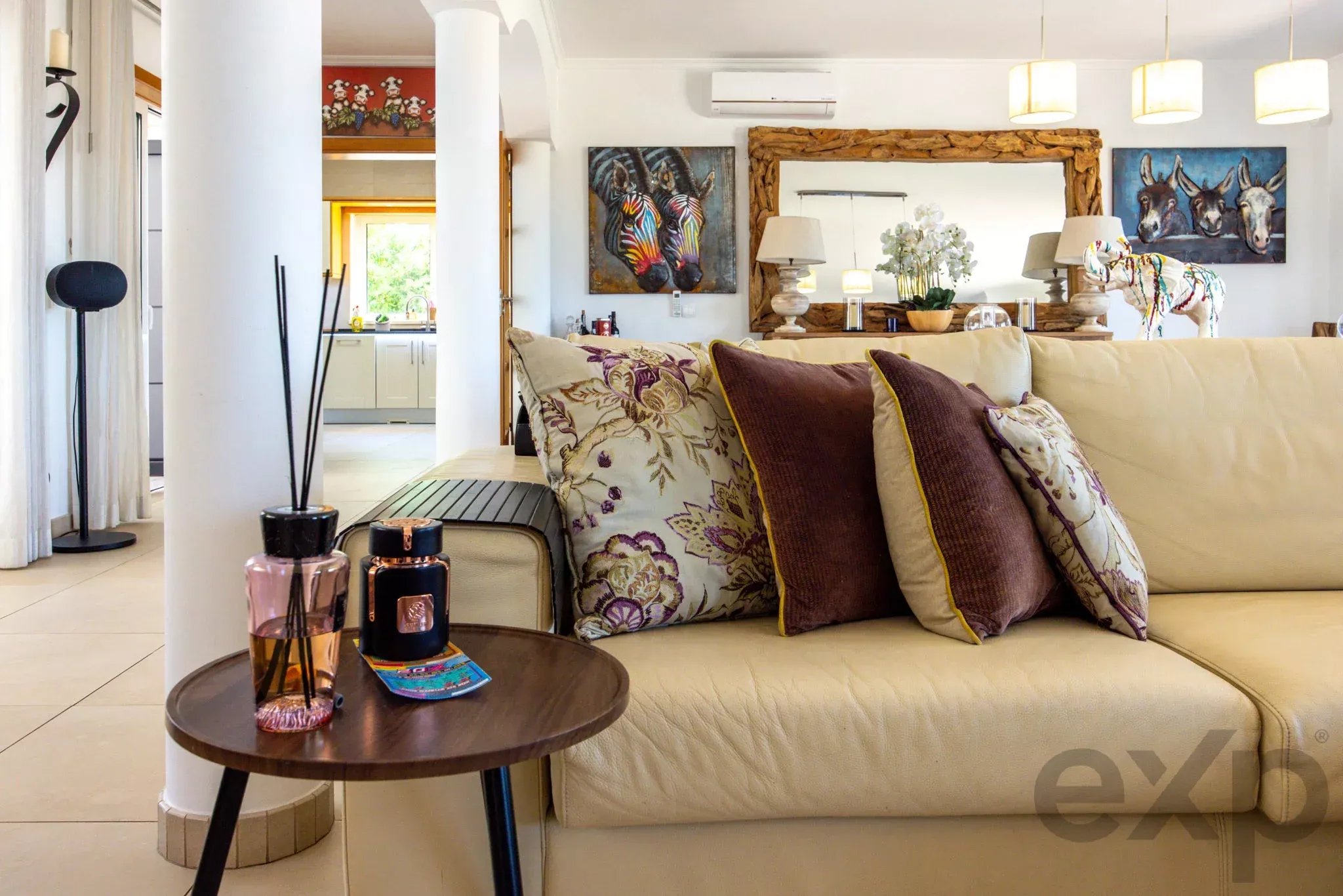 Living room with a beige sectional, floral cushions, and a round wooden coffee table holding candles and bottles.