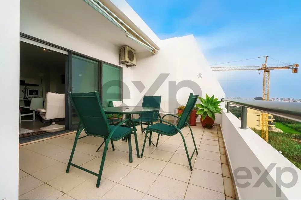Outdoor balcony with a glass-top table and four green chairs, white-tiled floor, and a potted plant, overlooking the city.