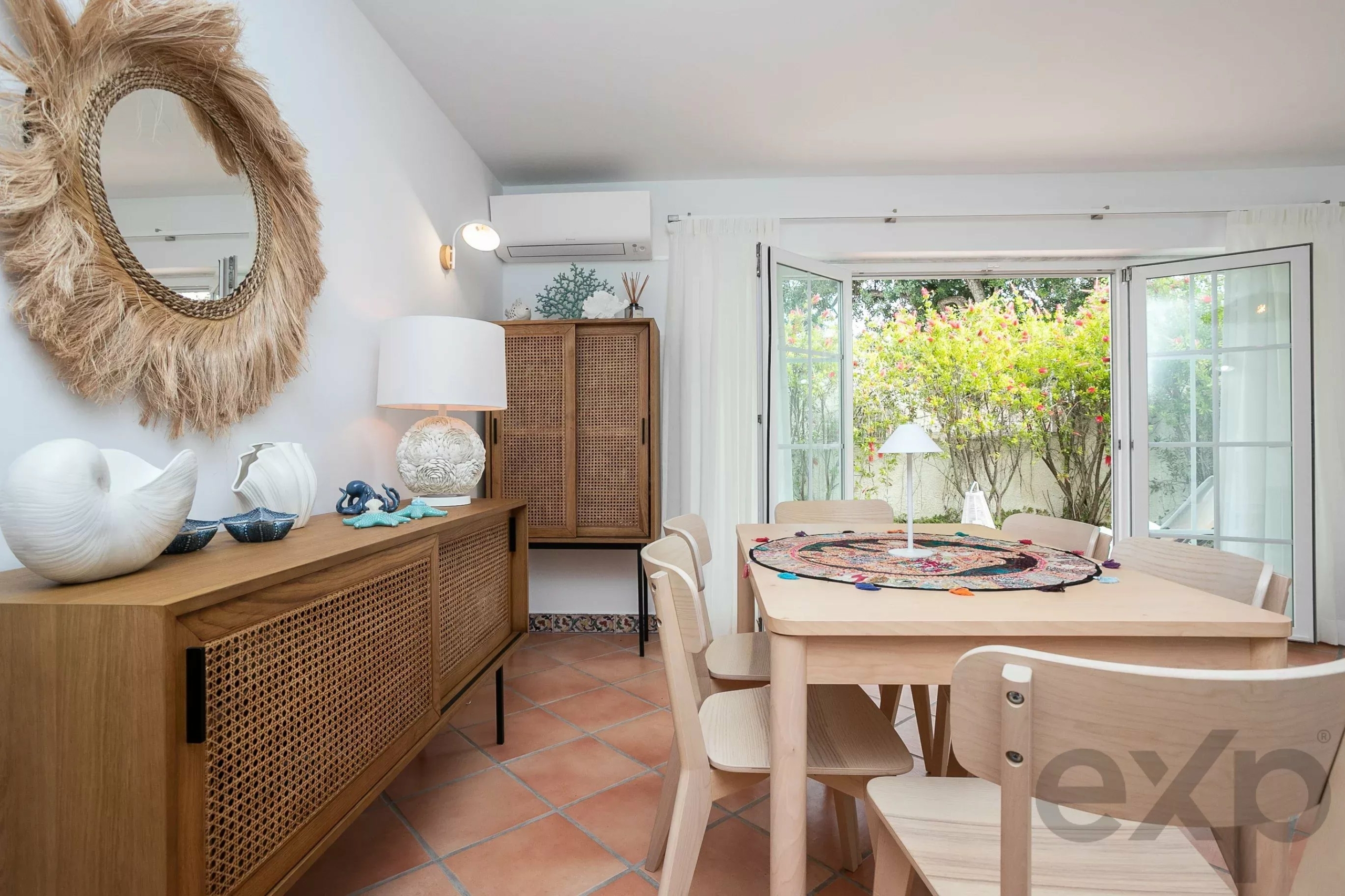 Round straw-framed mirror on a light wall above a wooden sideboard with wicker panels, tropical decor nearby.