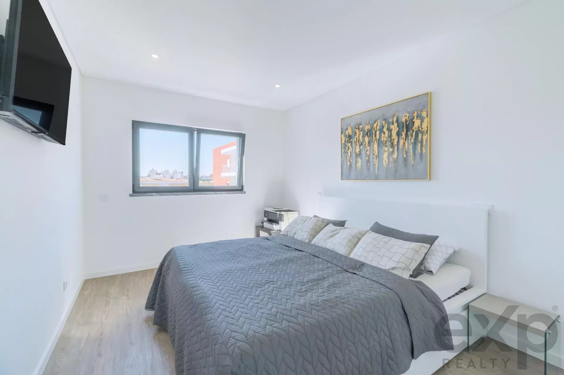 Modern white bedroom with a gray quilted bed, wall art, and a wall-mounted TV on the left.