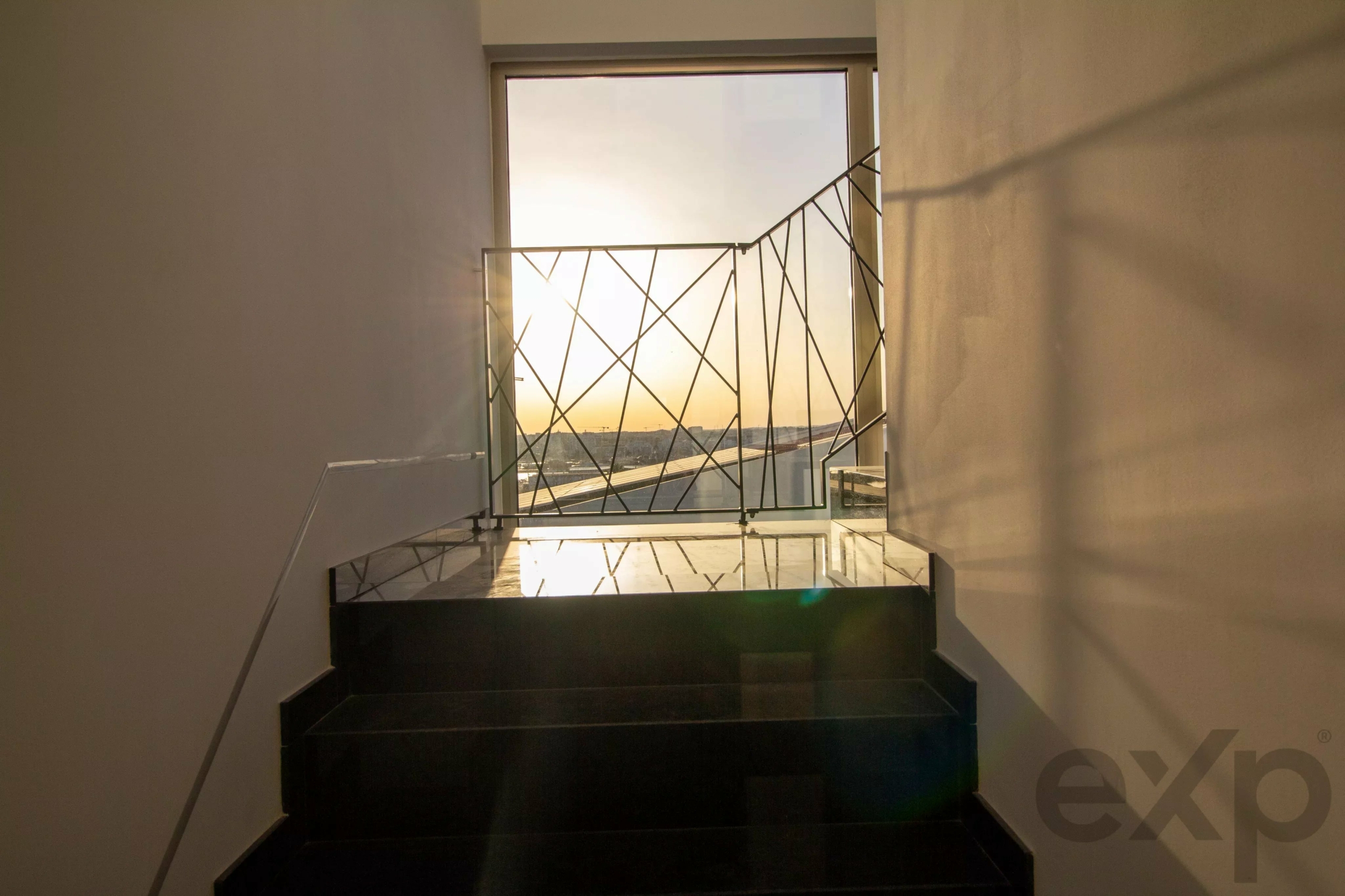 Indoor stairwell opening to a balcony with a geometric metal railing and sunset beyond the railing, seen from the stairs.