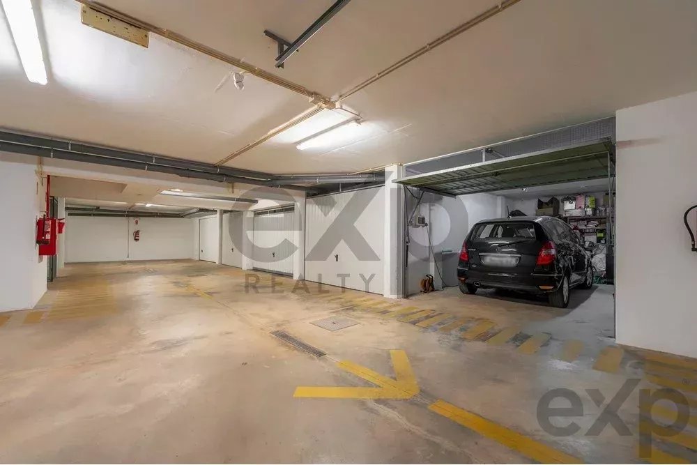 Underground parkinggarage with a car parked in a bay on the right, yellow directional arrows and parking lines on the concrete floor, and storage shelves nearby.