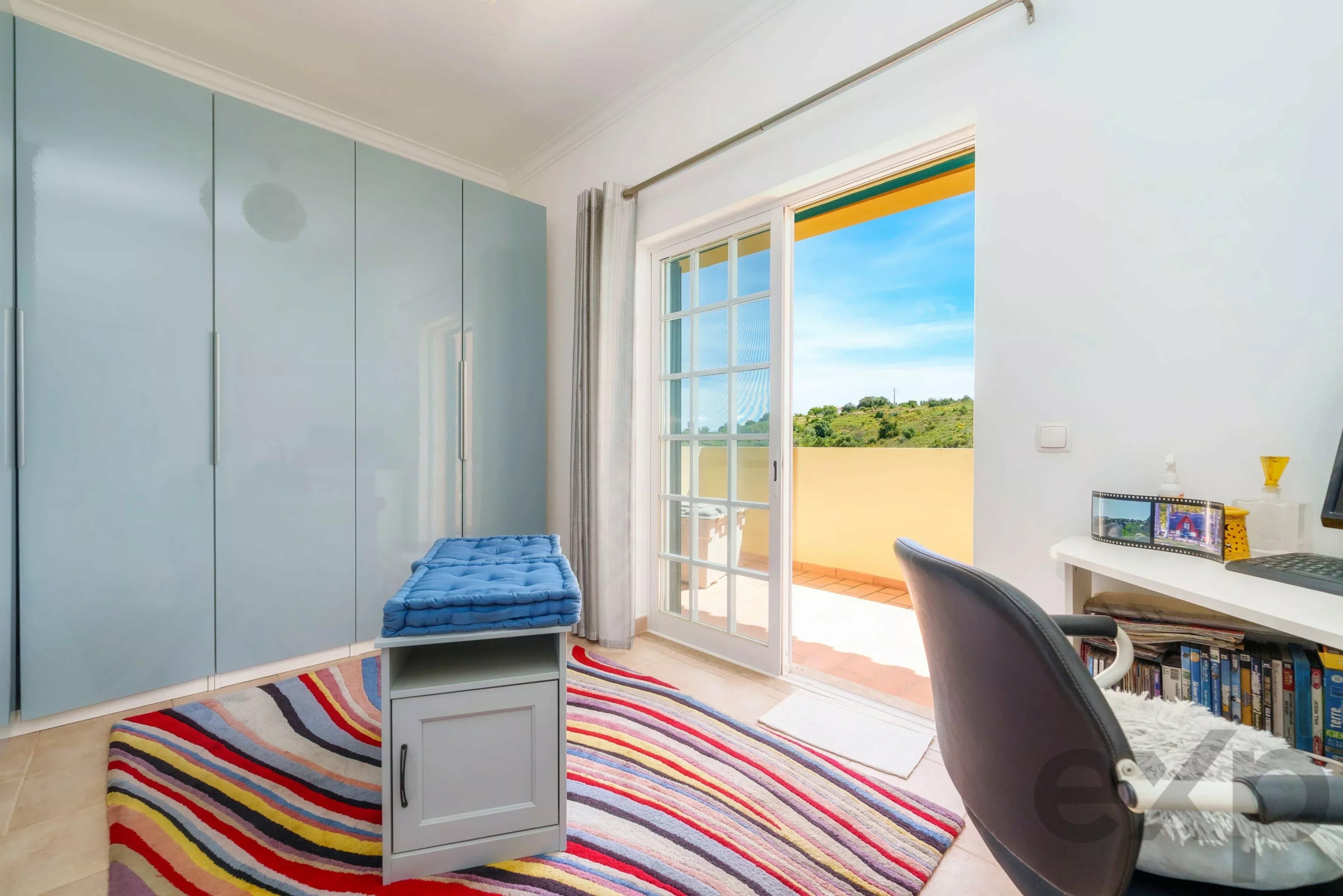 Bright room with light blue wardrobes along the left wall, a blue cushioned bench, and a multicolored striped rug.