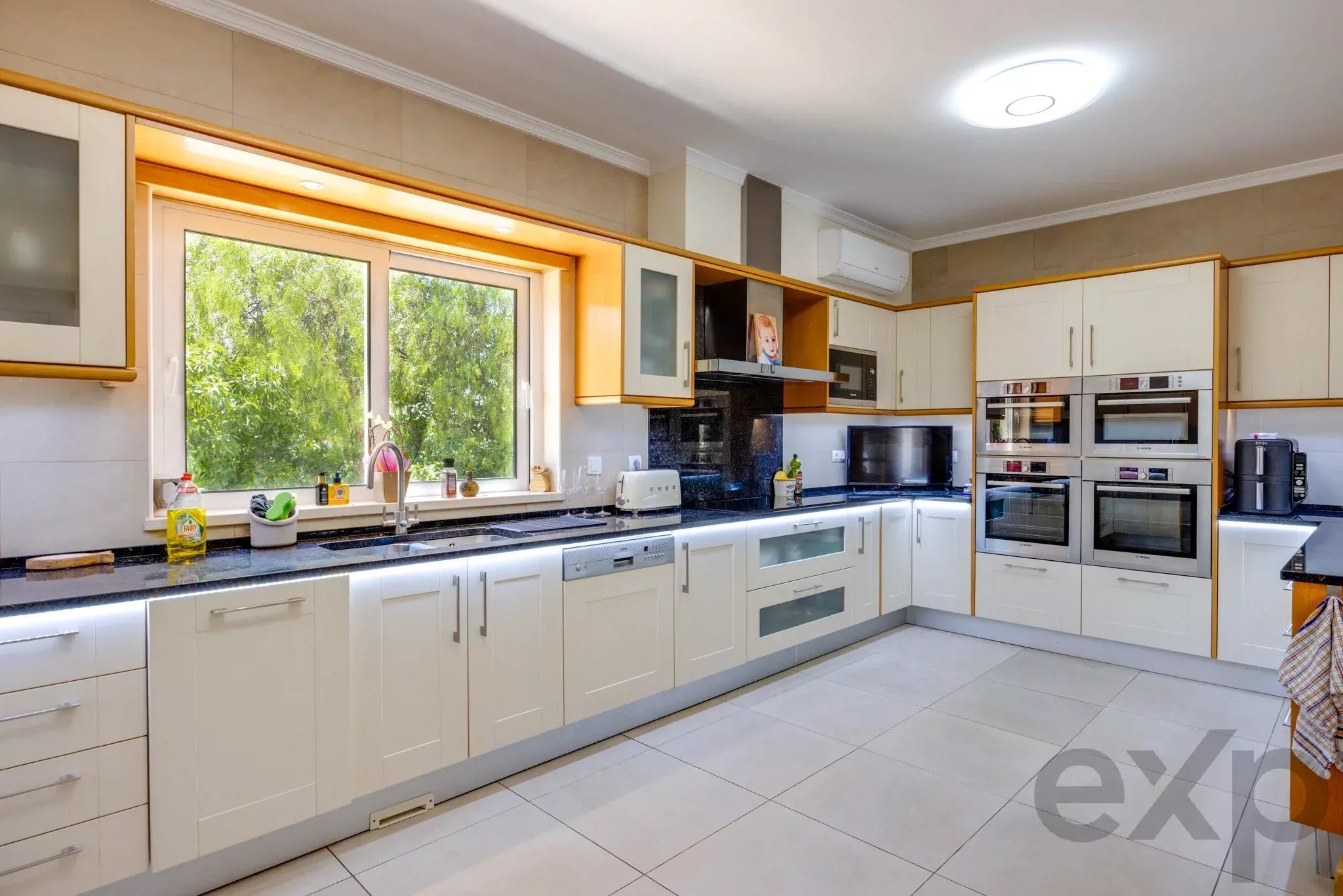 Bright modern kitchen with white cabinets, black countertops, and under-cabinet lighting; large window view of greenery on the left.