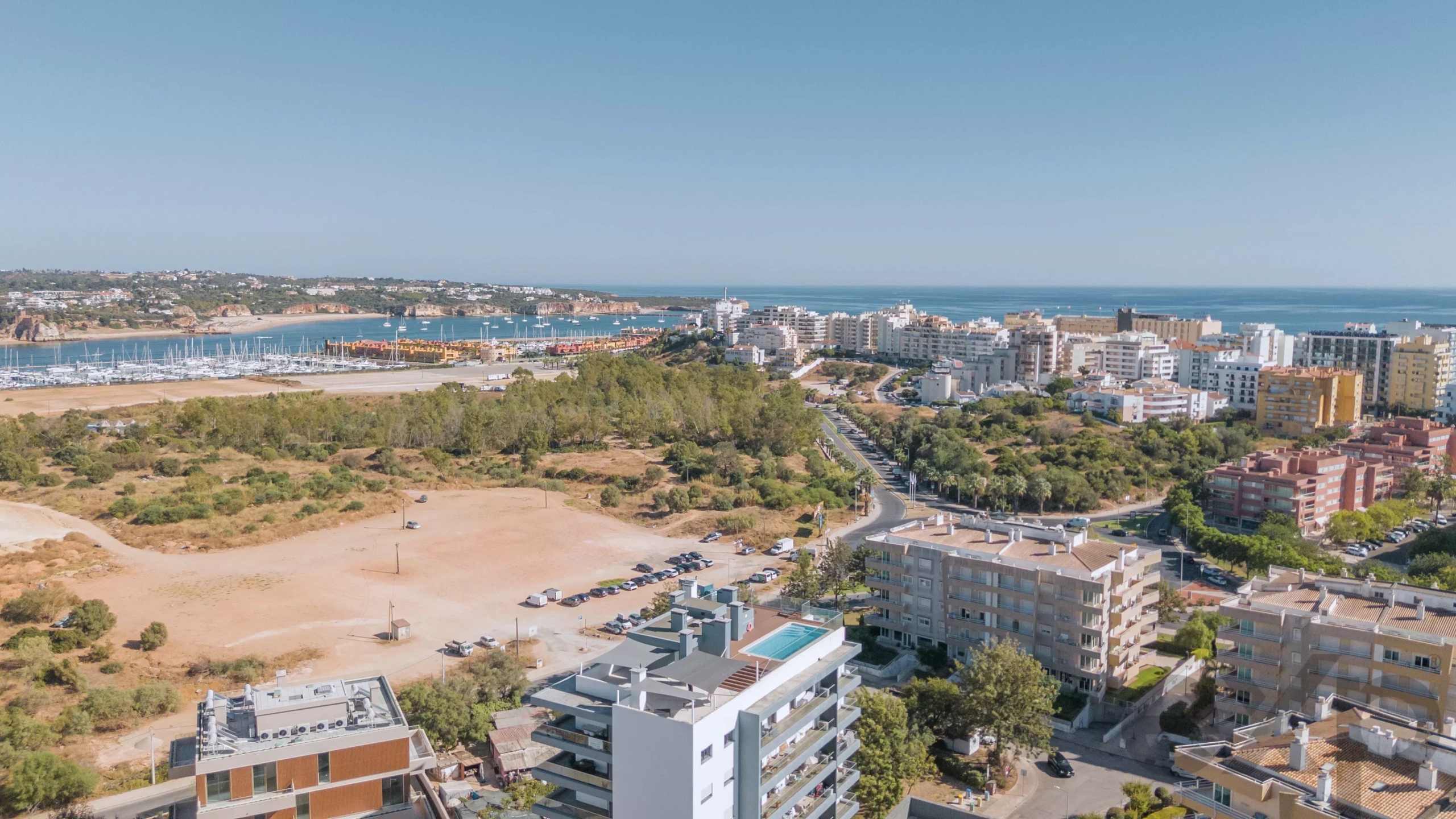 Aerial view of a seaside city: marina with sailboats, white apartment blocks along the shore, and green hills in the foreground.