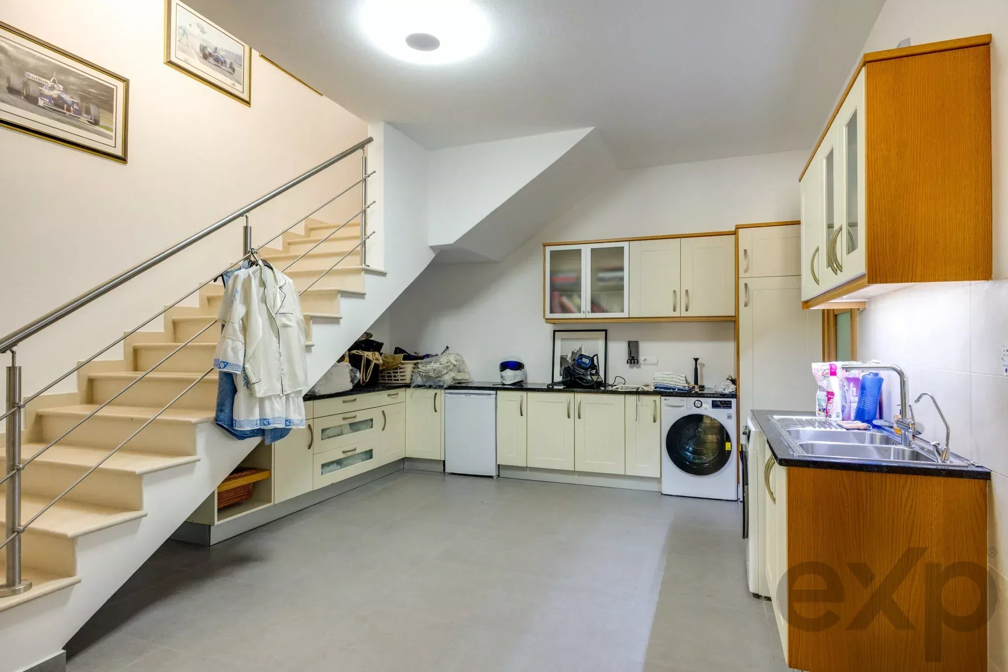 Kitchen under a staircase with white cabinets, washing machine, and sink; stainless steel railing along stairs on the left.
