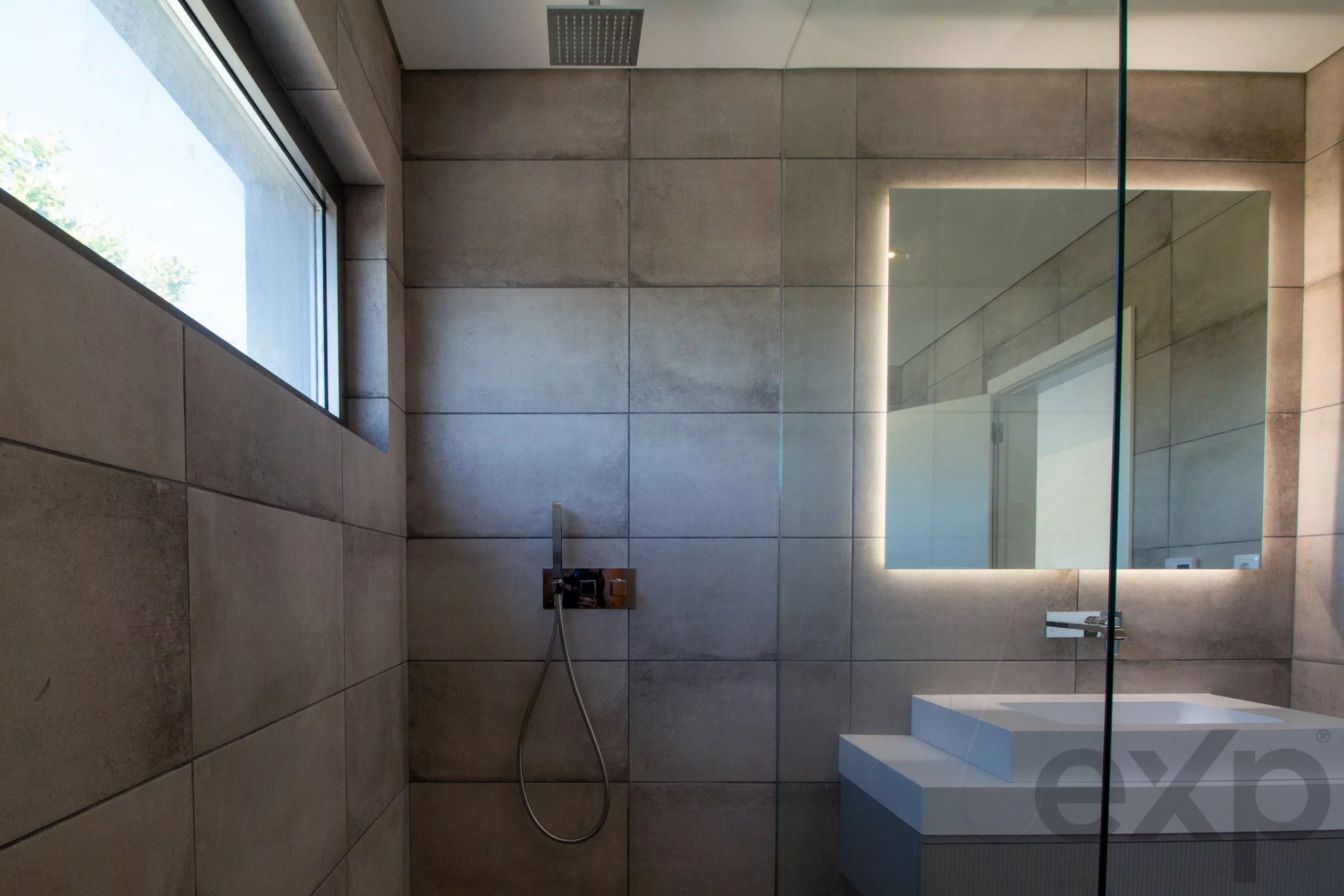 Modern bathroom with large grey tiled walls, a square overhead shower head, glass divider, backlit mirror, and a white sink counter visible on the right.