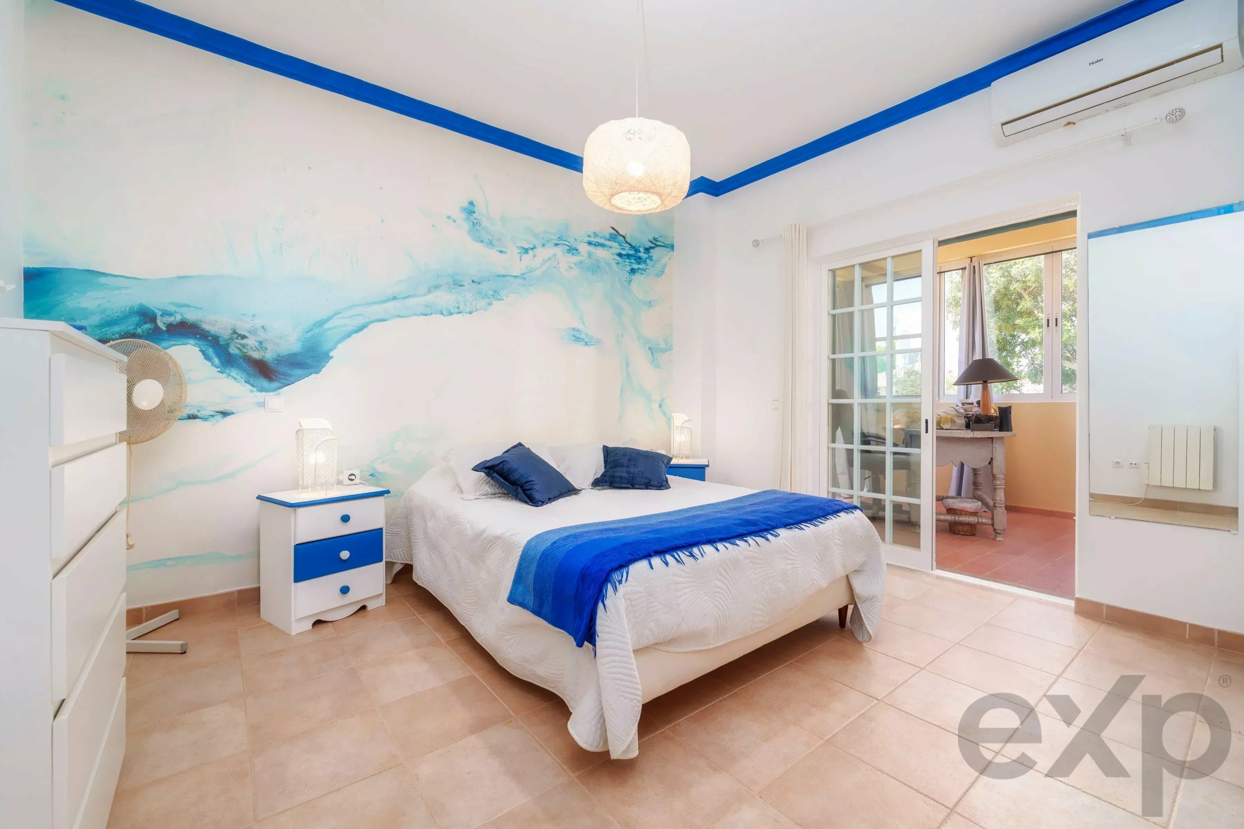 Bright coastal-themed bedroom with white furniture, blue accents, and a wall mural of blue waves offering a seaside vibe.