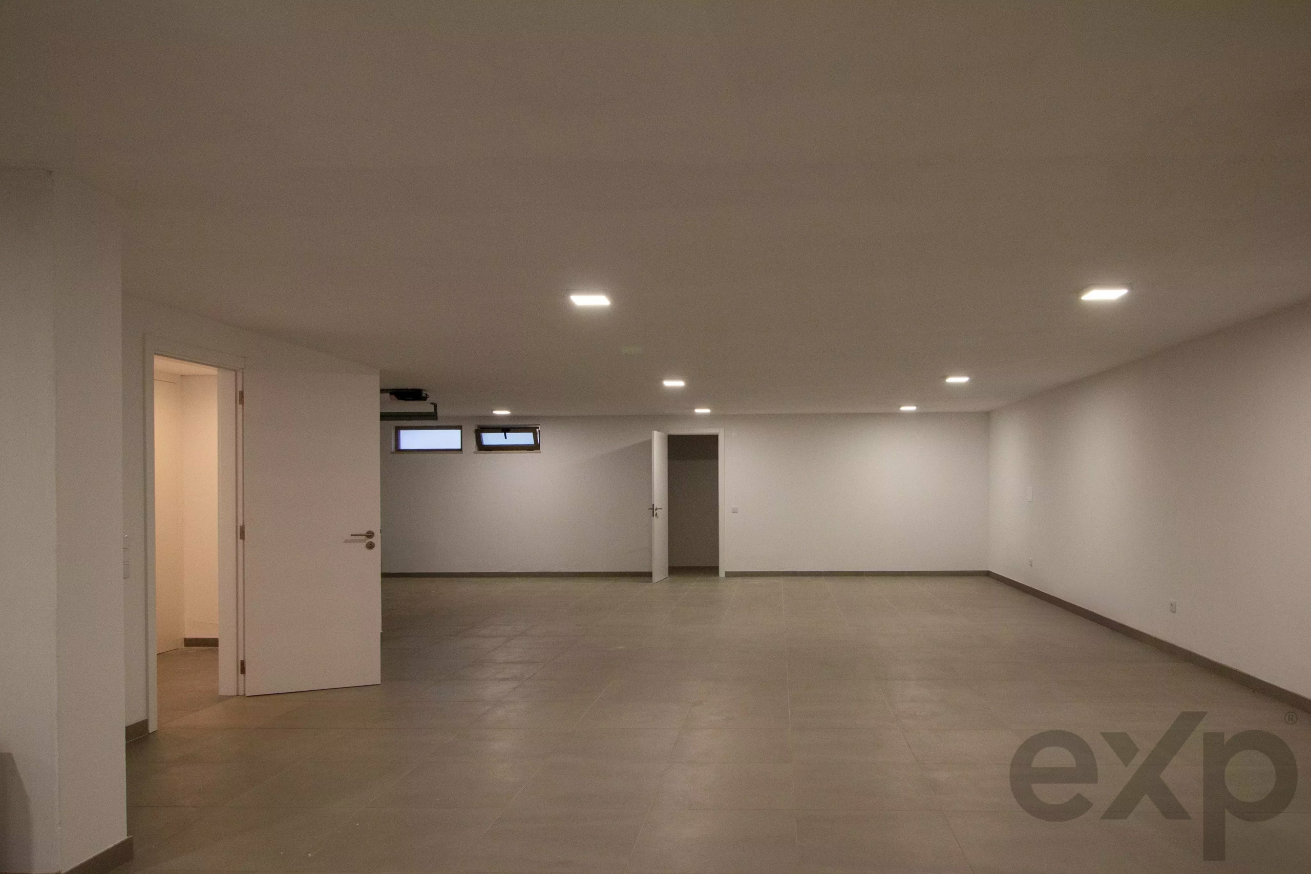 Spacious empty office area with white walls, gray tiled floor, and recessed ceiling lights.