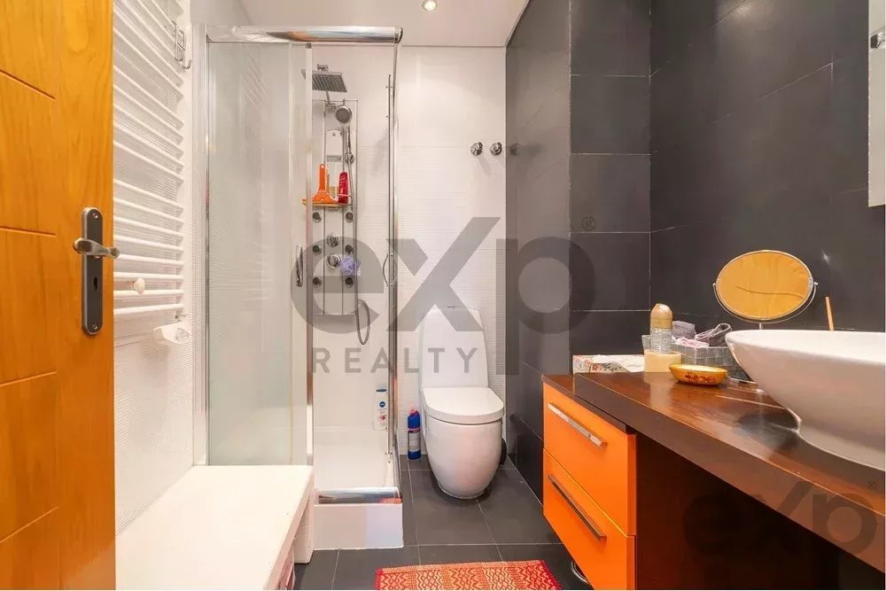 Modern bathroom with a glass-enclosed shower, white toilet, dark tiled walls, and an orange-drawer wooden vanity with a round vessel sink.