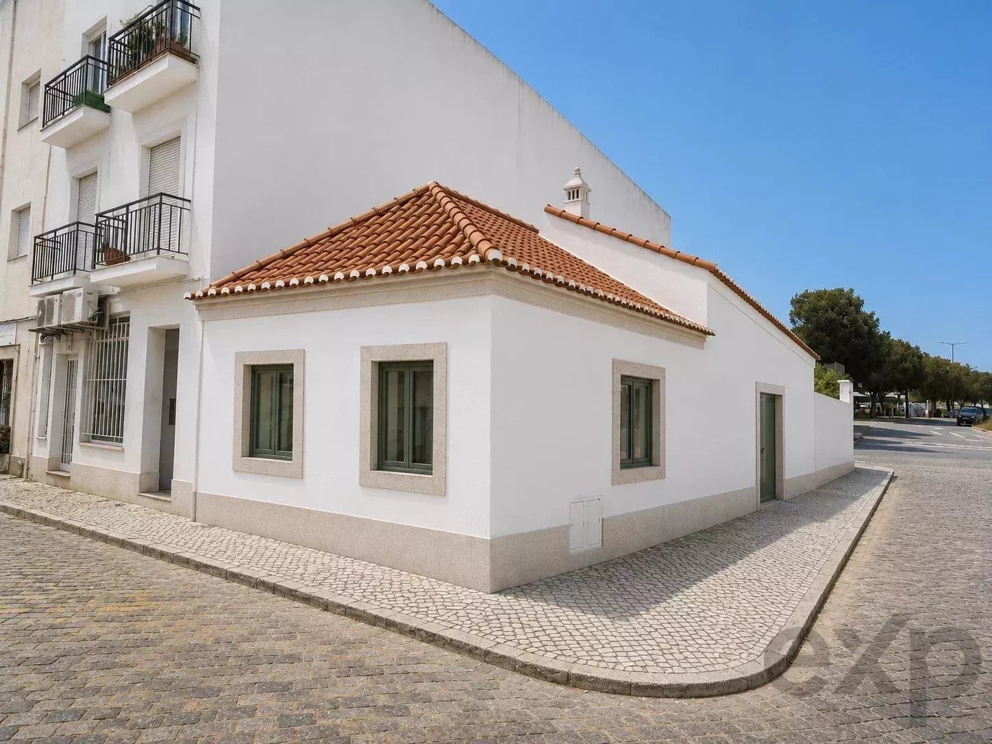 White single-story house with a red-tiled roof on a cobblestone street, at a street corner under a blue sky.