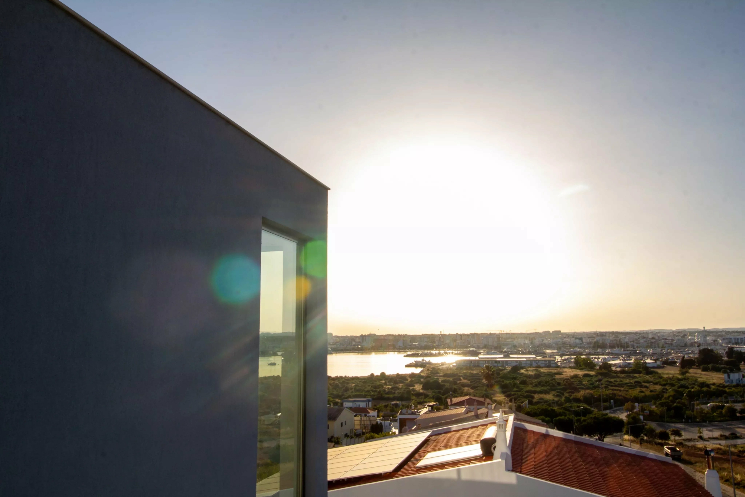 Sunlit cityscape seen from a balcony, with a dark wall on the left and the bright sunset over water in the distance.