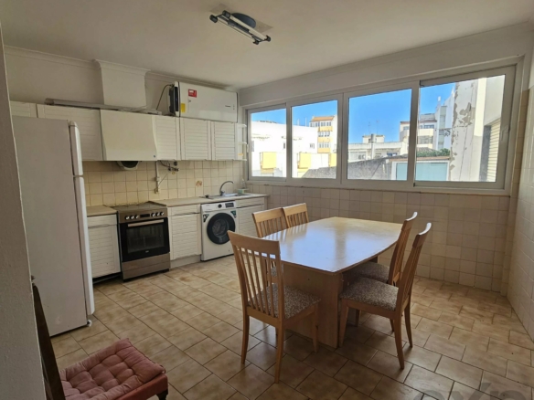 Bright kitchen with white cabinets, a table and wooden chairs by a windowed wall, appliances on the left, and daylight outside