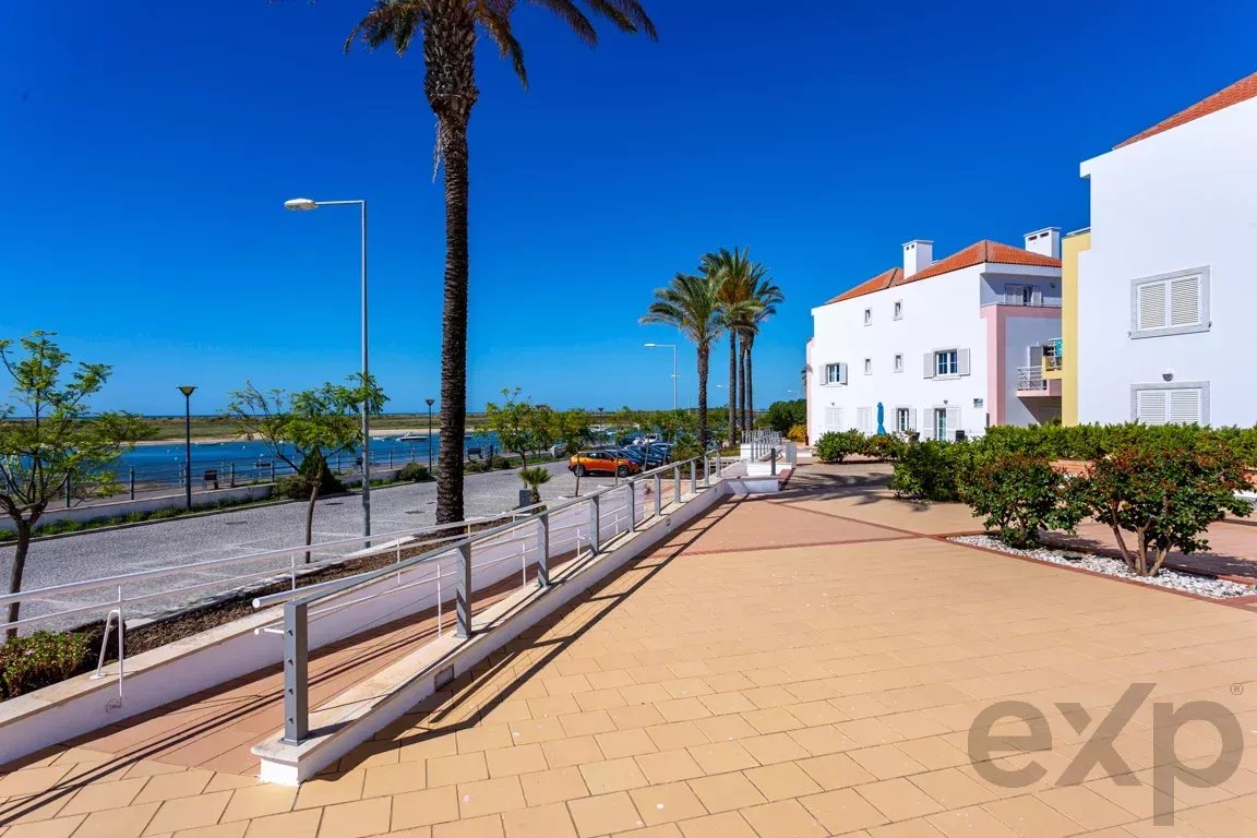 Sunlit coastal promenade with palm trees, white buildings, and a deep blue sky by the sea.