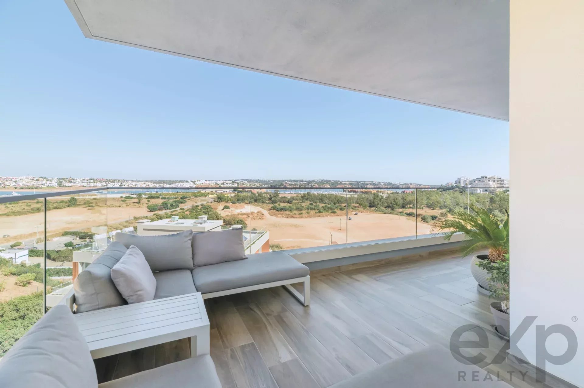 Modern balcony with a pale gray outdoor sofa, white coffee table, and glass railing overlooking a sandy landscape and distant town under a clear blue sky.