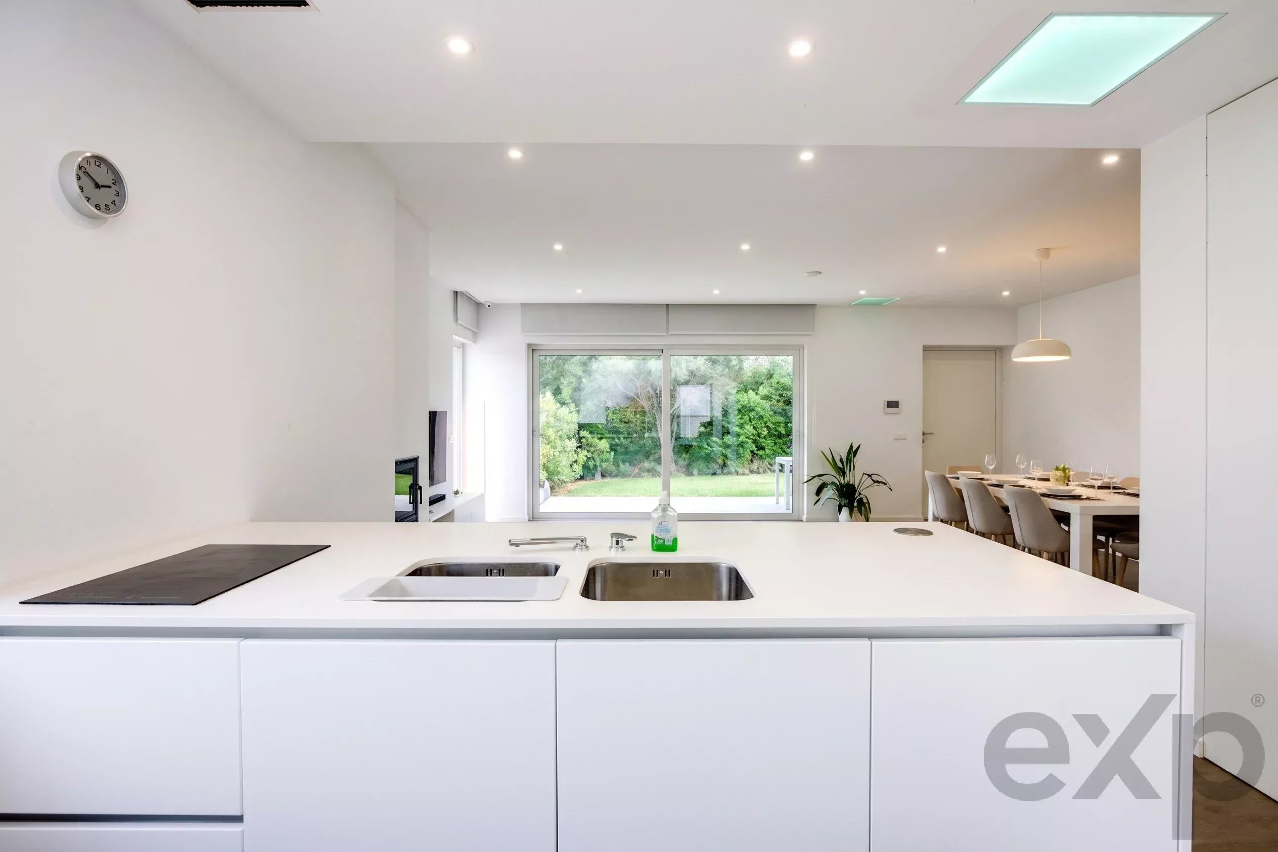 Bright white kitchen with a central island, double sink, and a dining area visible in the background.