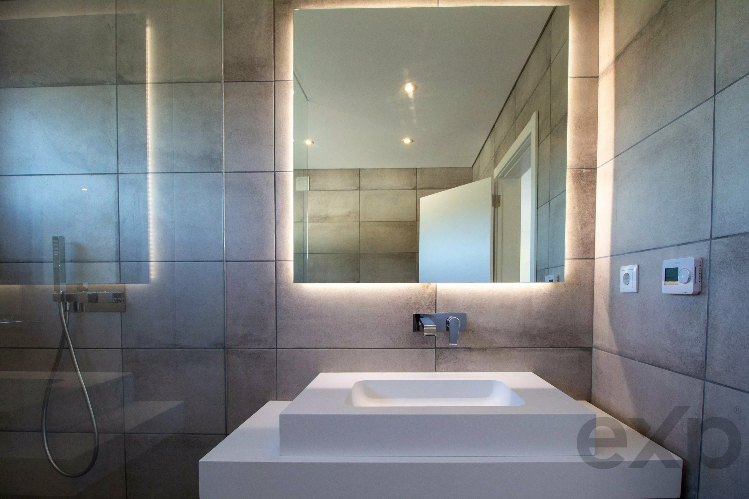 Modern bathroom with a backlit rectangular mirror above a white rectangular sink and wall-mounted faucet; gray tiled walls and a glass shower panel on the left.