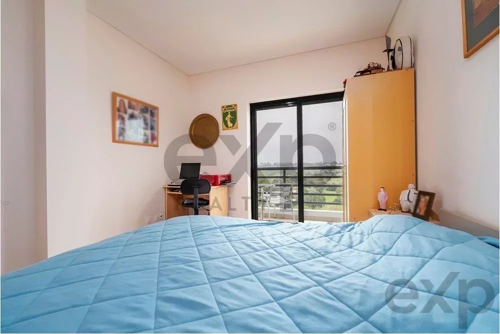 Bedroom with a blue quilted bed in the foreground, a desk and chair near the wall, and balcony doors at the far end.