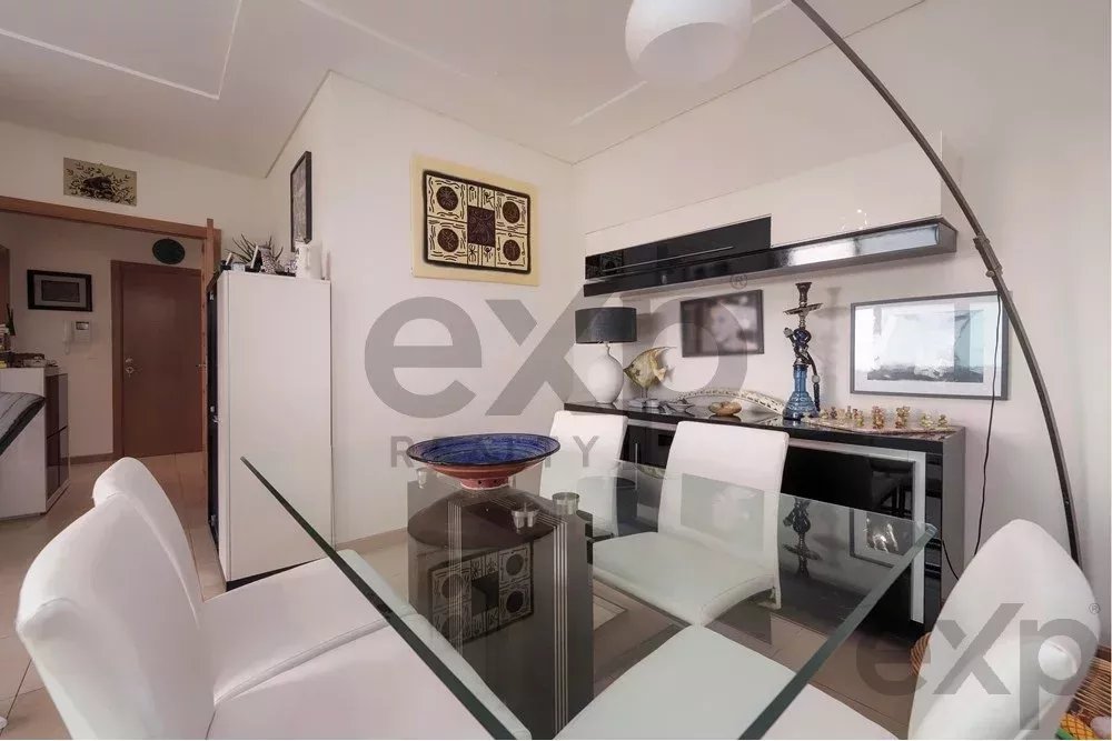 Bright, modern dining room with a glass table, white chairs, and a kitchen visible in the background.