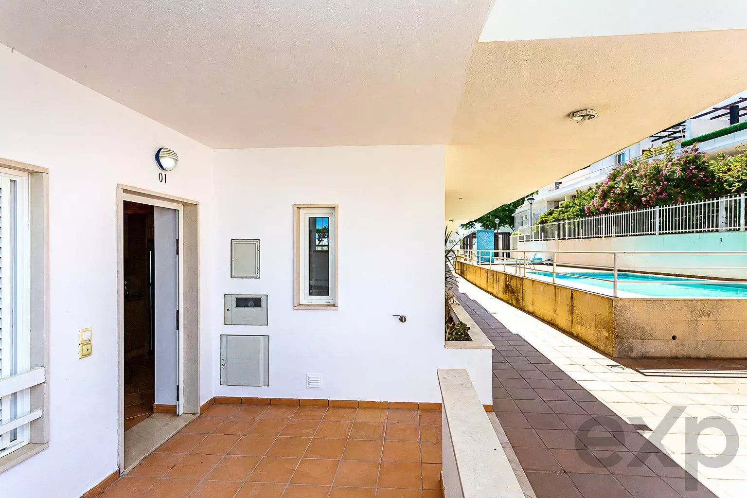 White apartment balcony with terracotta floor tiles, open door, small window, and utility panels; pool area visible along a walkway to the right.