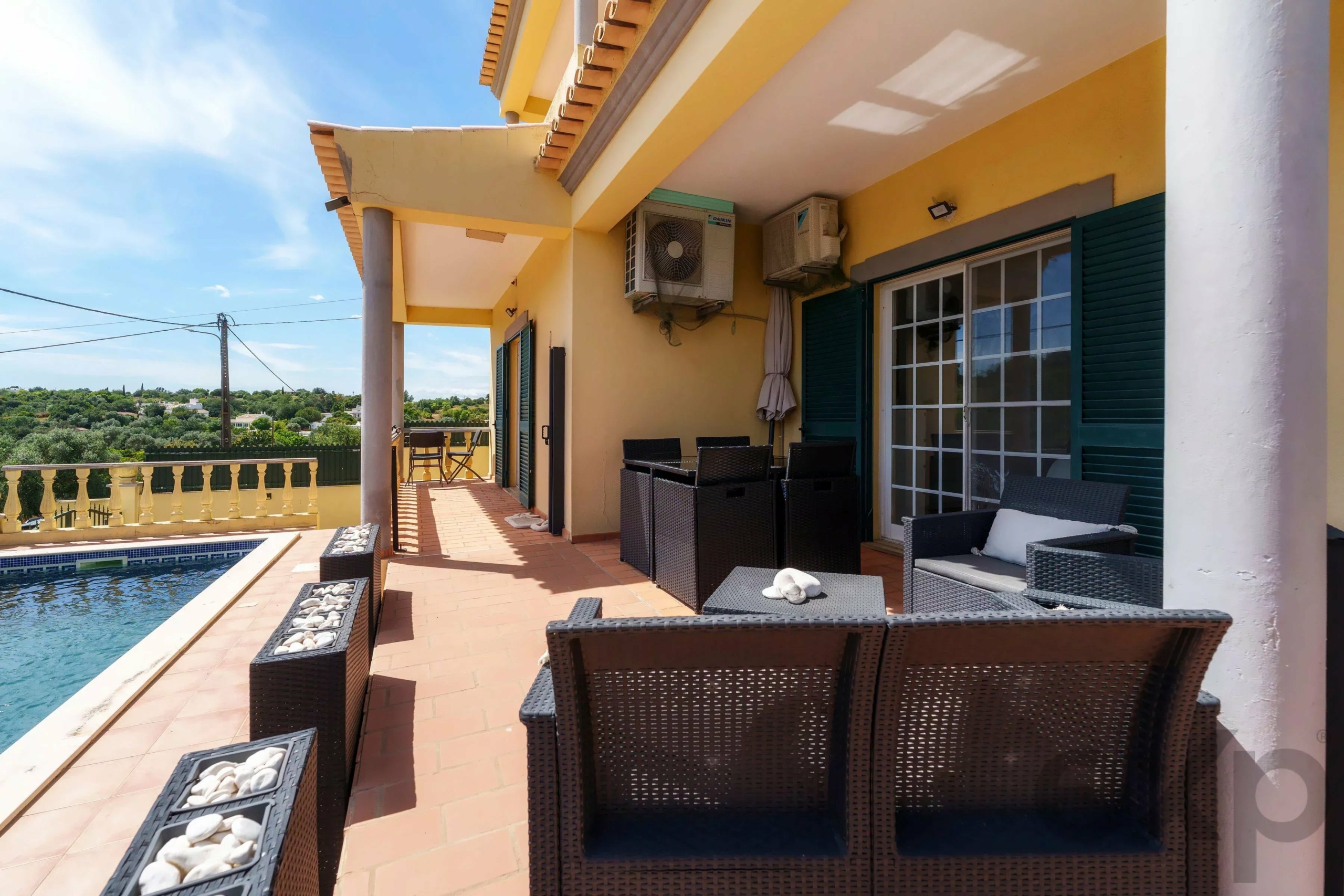 Sunny terrace with wicker seating and a pool to the left, yellow building walls, and clear blue sky above.