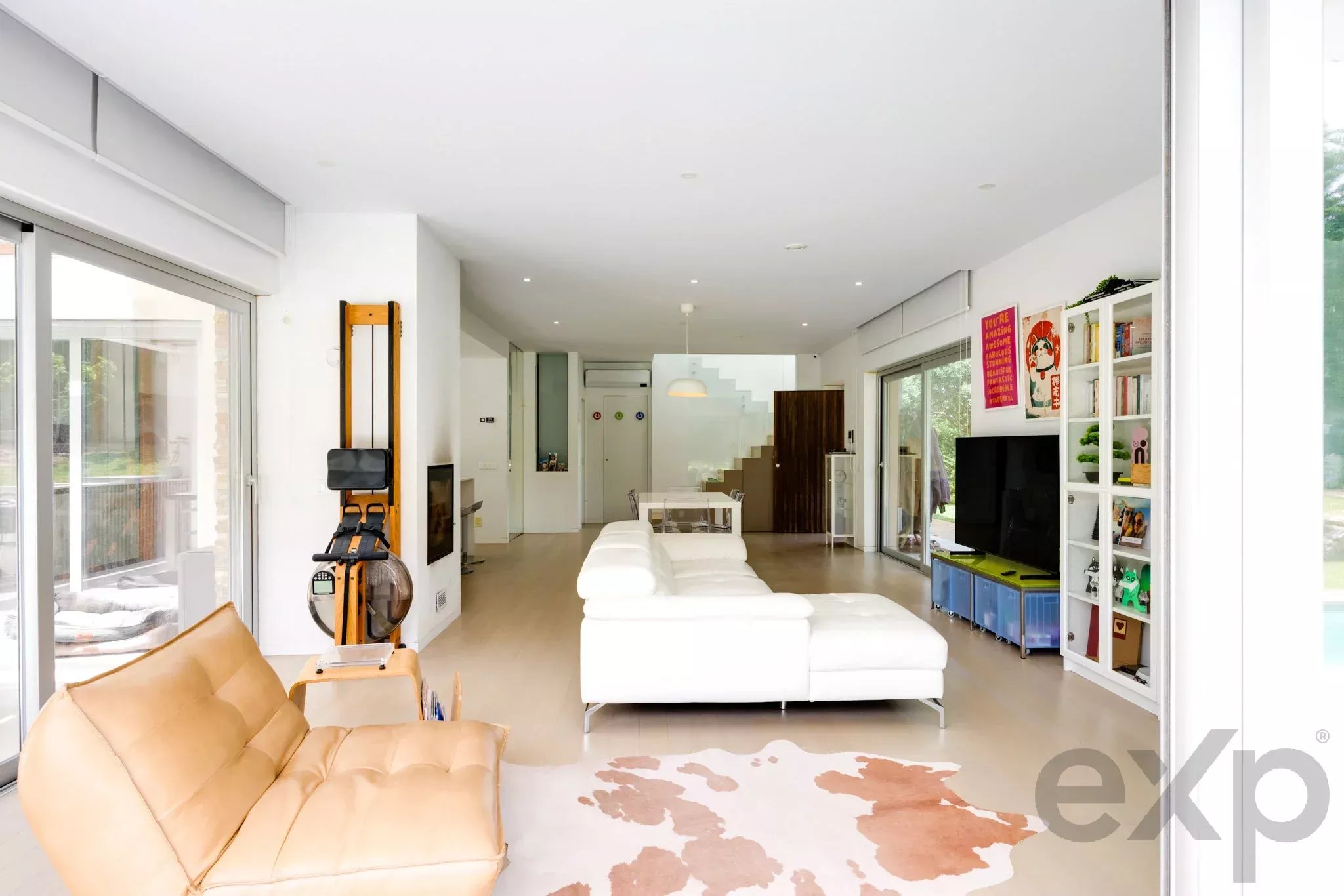 Open, bright living room with white sofa set, beige leather couch, and a glass sliding door wall letting in natural light; a white bookshelf and TV on the right, and a staircase in the back. A piece of home gym equipment stands near the left wall.