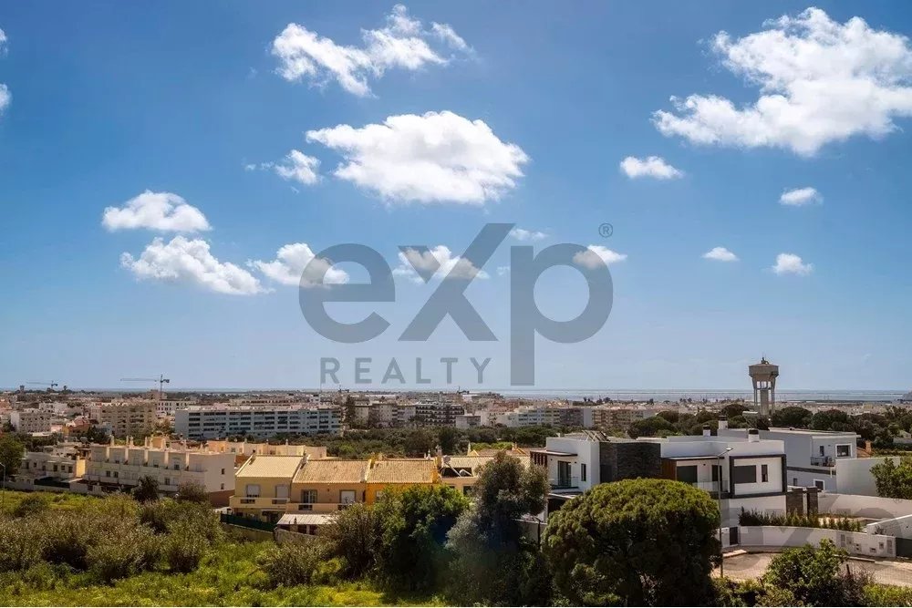 Coastal city skyline with low-rise houses, greenery in the foreground, and the sea on the horizon under a blue sky with scattered clouds.