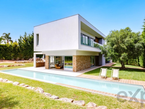Modern two-story white house with glass balcony overlooking a rectangular pool and grassy yard beneath a clear blue sky, surrounded by trees.