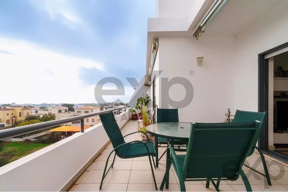 Balcony with a round glass table and green chairs on a tiled terrace, overlooking a residential neighborhood under a blue sky.