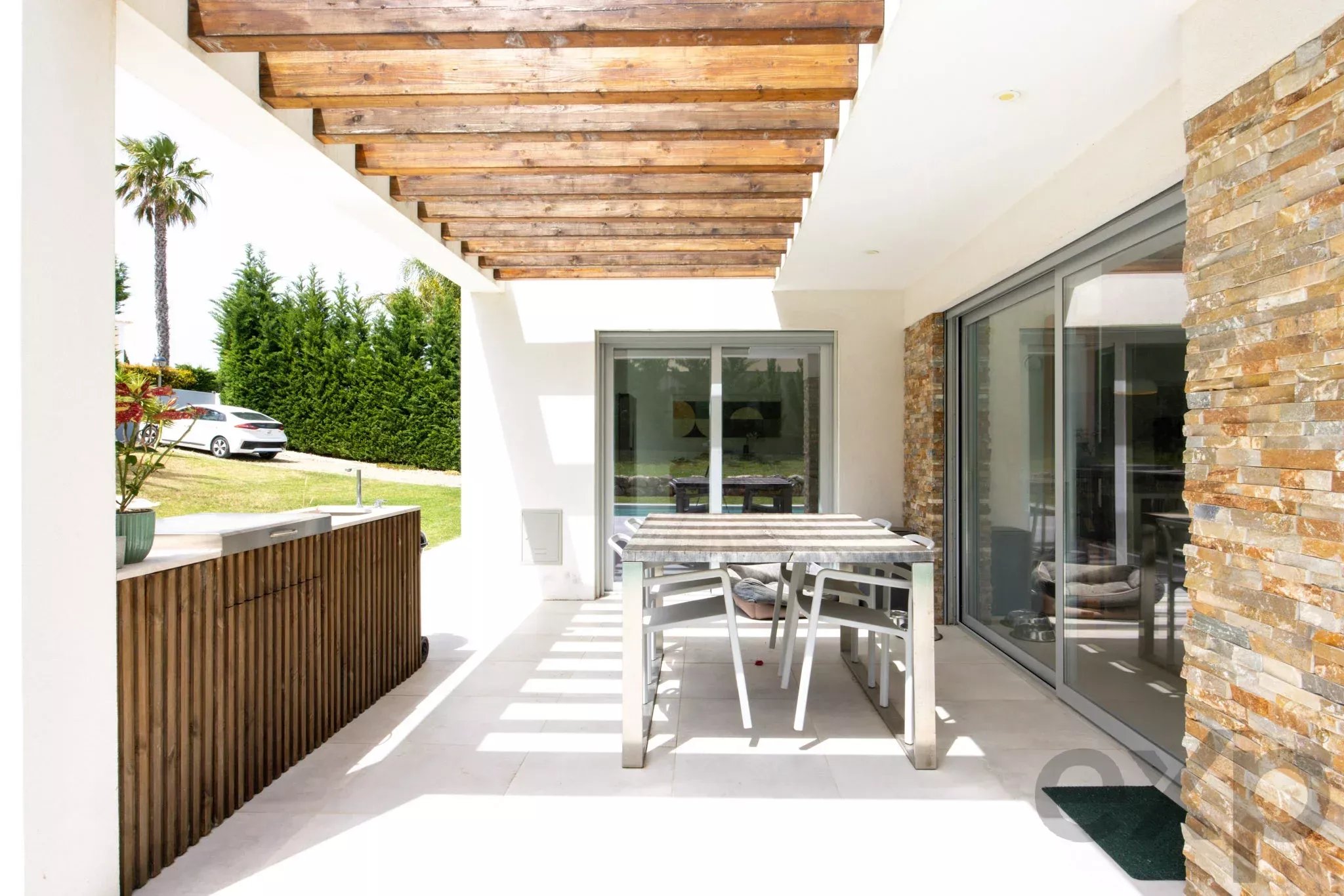 Outdoor patio with a wooden pergola, white walls, and a glass sliding door; a dining table and chairs sit on a light tiled floor beside a stone wall and hedge.”