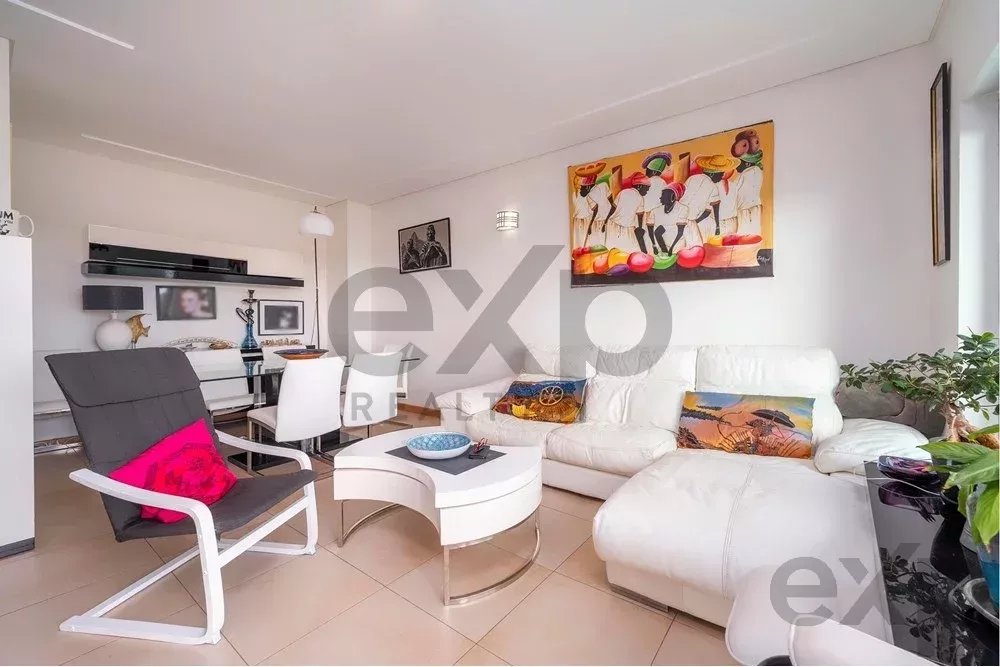 Bright contemporary living room with a white sectional sofa, black lounge chair, glass coffee table, and colorful wall art; plants and decorative objects add color.