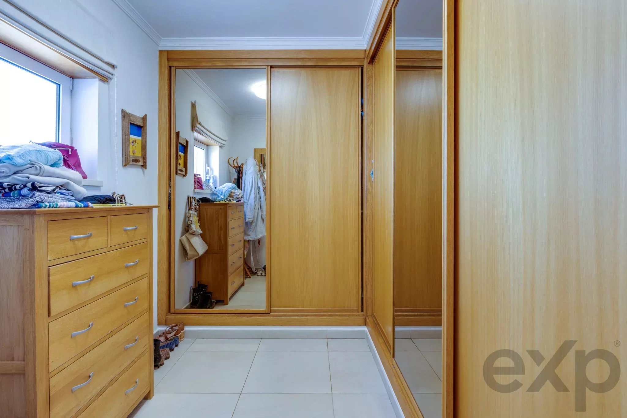 Walk-in closet with light wooden wardrobes, a mirrored sliding door, and piles of folded clothes on a dresser; shoes on the tiled floor.
