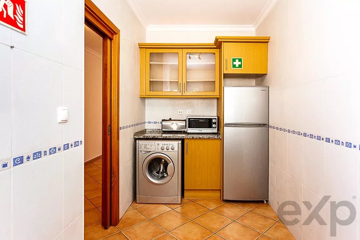 Compact kitchen with a washing machine under the counter, a tall stainless fridge, microwave and toaster on a granite countertop, and wooden cabinets above.