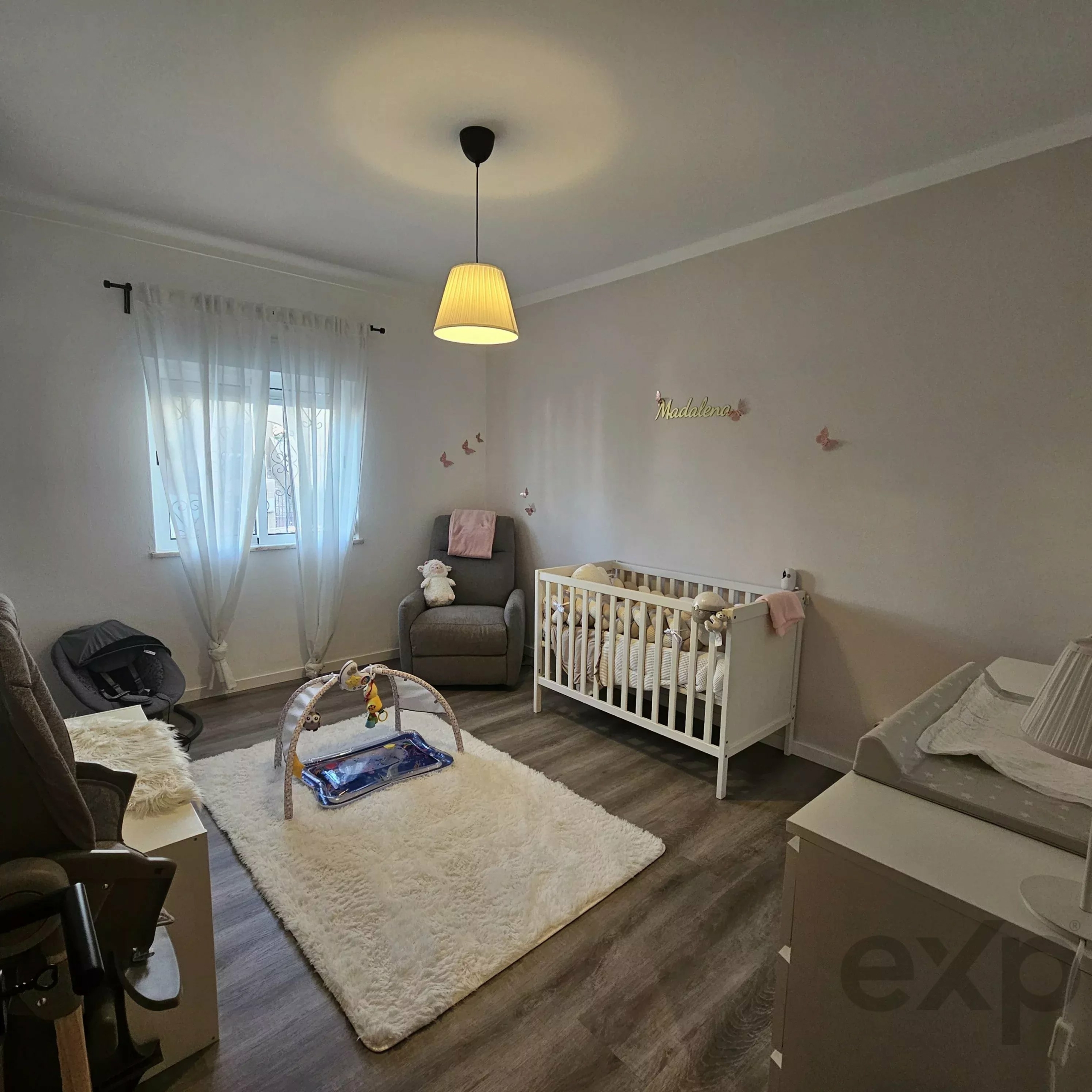 Nursery with a white crib, gray glider chair, and a fluffy white rug; sheer curtains and butterfly wall decals.