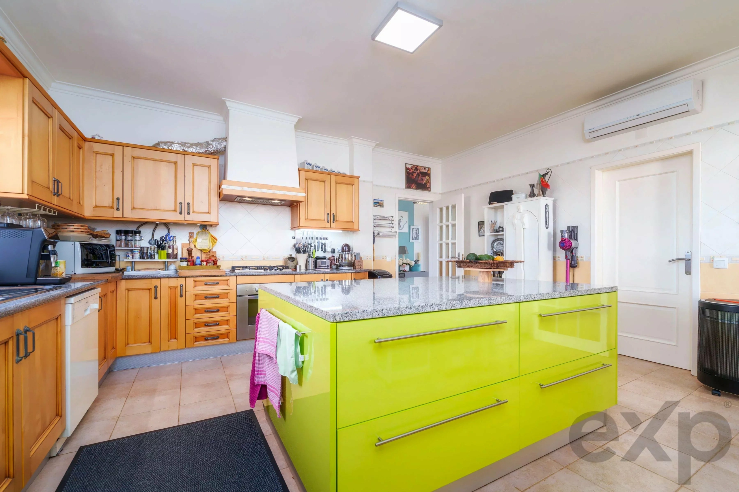 Bright kitchen with lime-green island center, wooden cabinets along the walls, and stainless steel appliances; a white-tiled backsplash and open shelving visible on the left.
