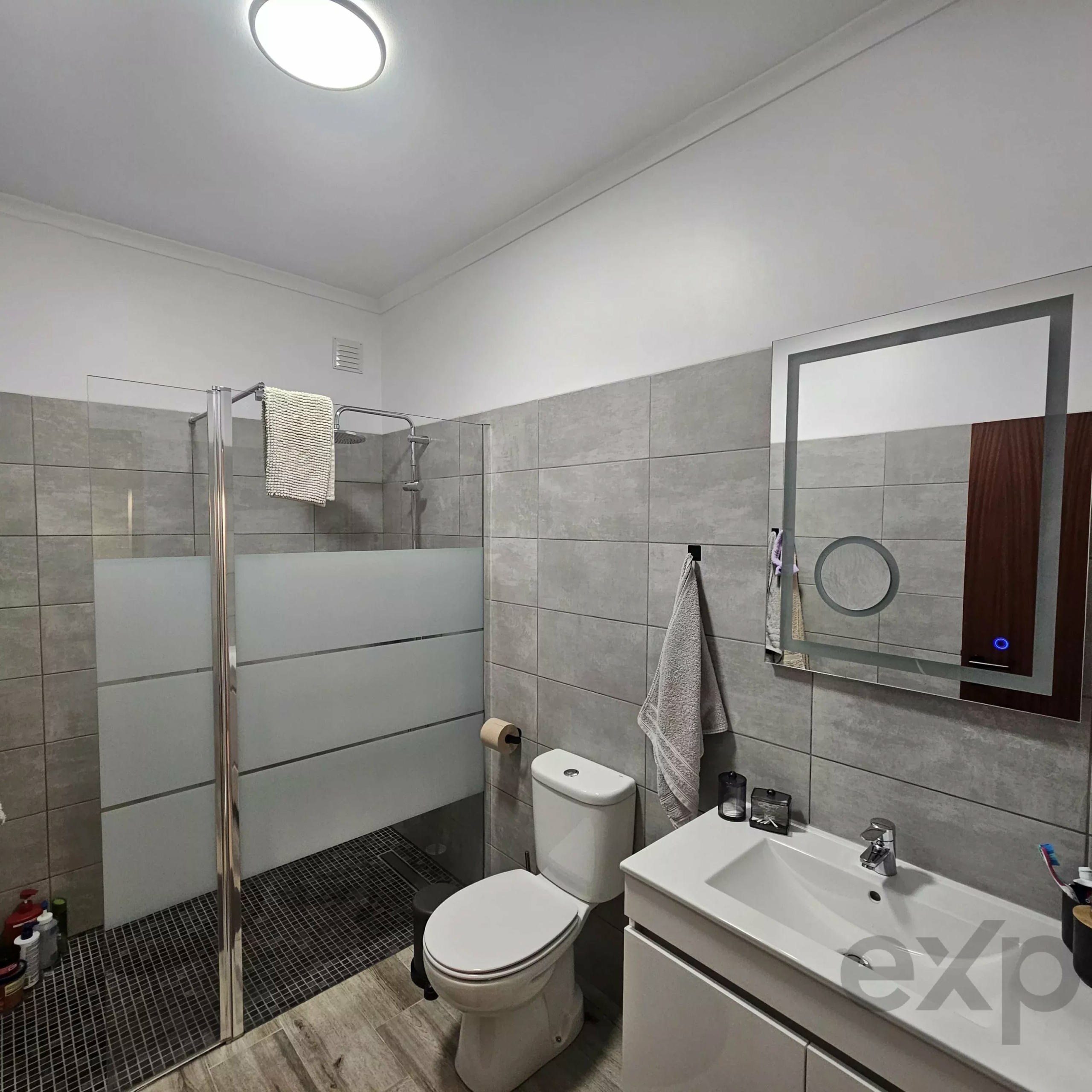 Modern bathroom with gray tile walls, glass shower enclosure, toilet, and white vanity with towels on a rack nearby.