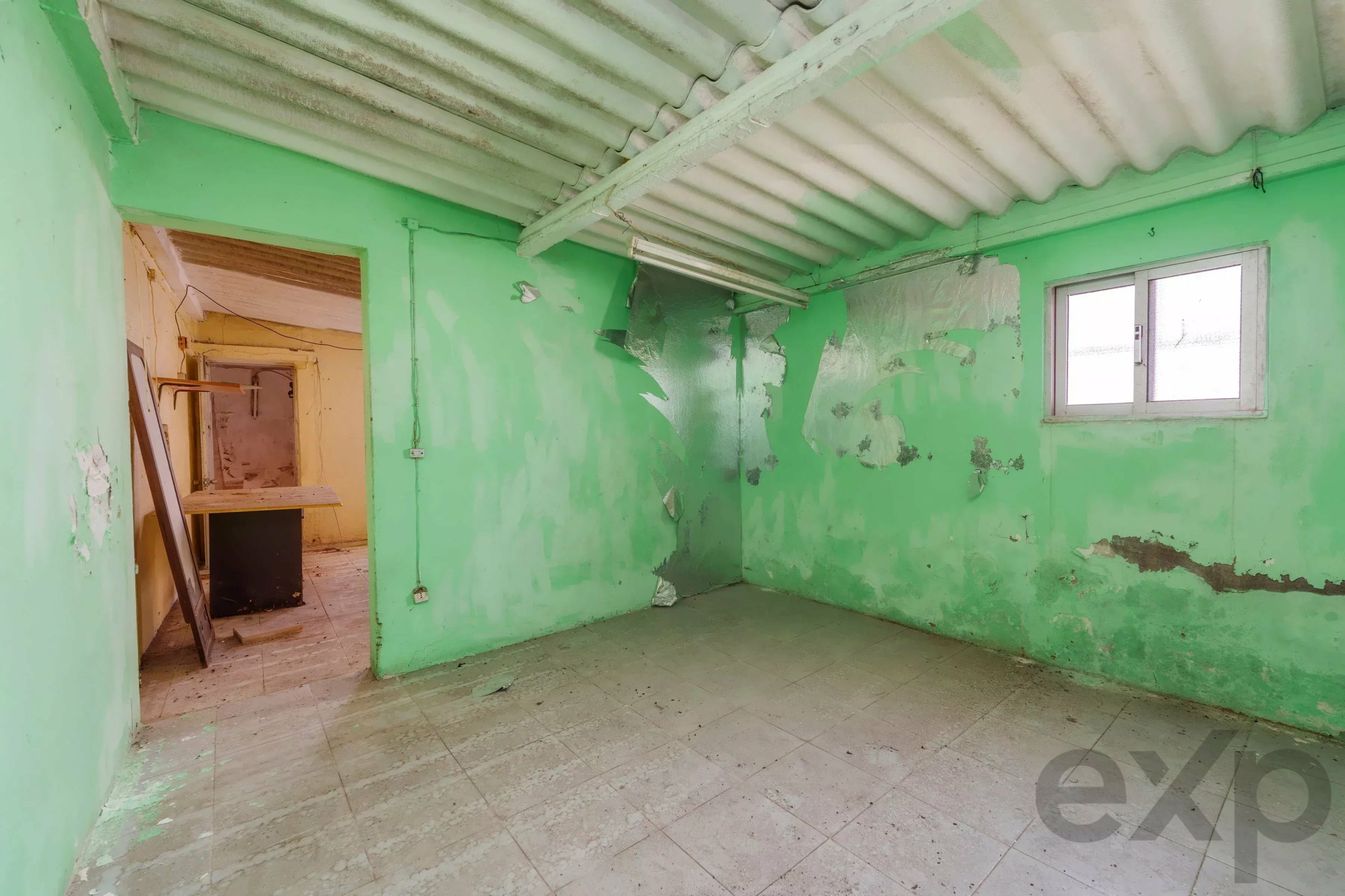 Abandoned room with green walls, peeling paint, and exposed ceiling beams; a small window on the right side.