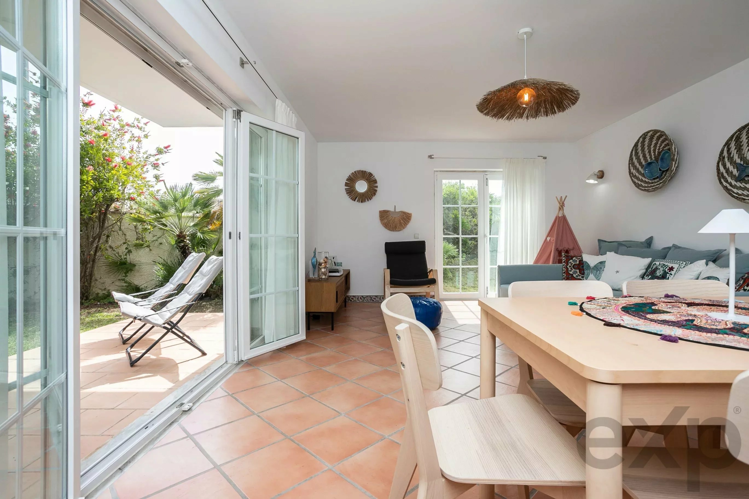 Bright living room with open French doors to a sunny patio and greenery outside, white walls, and coastal decor including a woven sunburst mirror and rope baskets on the wall.