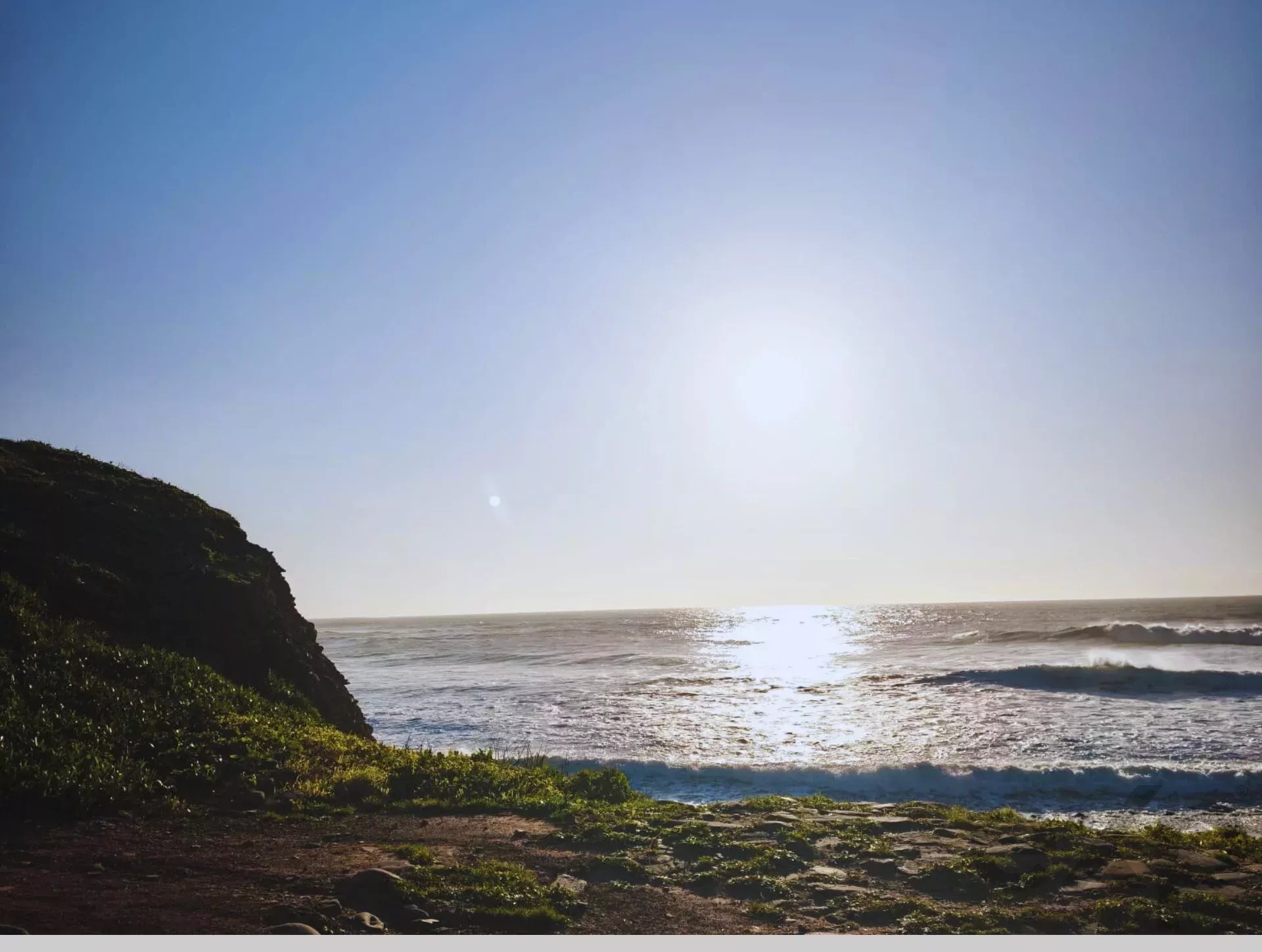 Sunlit coastal landscape: rocky cliffside on the left, calm ocean and a bright sun shining over the water.
