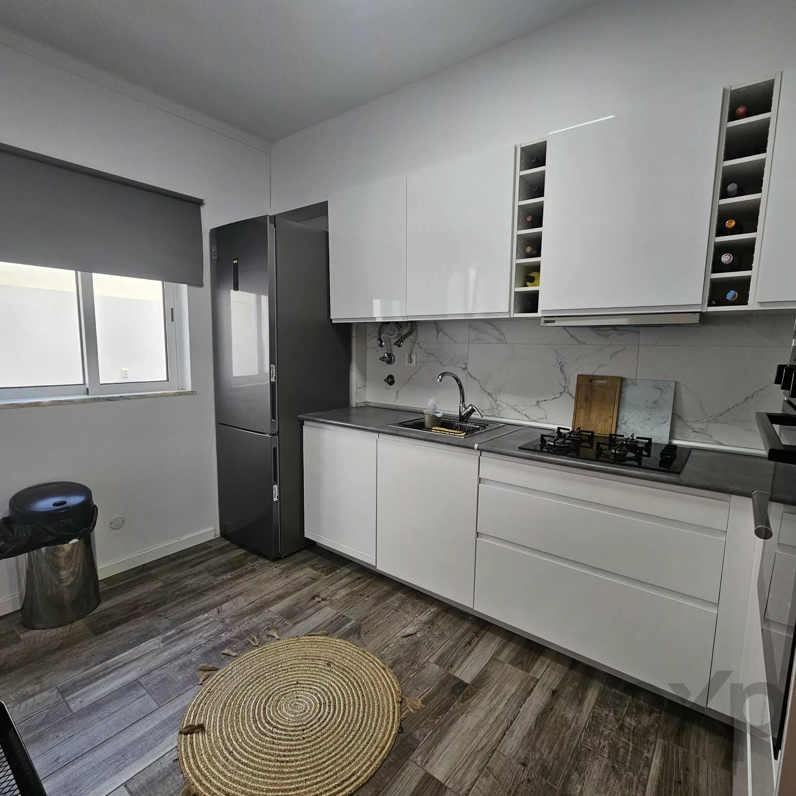 Modern kitchen with white high-gloss cabinets, a stainless steel fridge, marble backsplash, and two circular woven rugs on a wood-like floor.