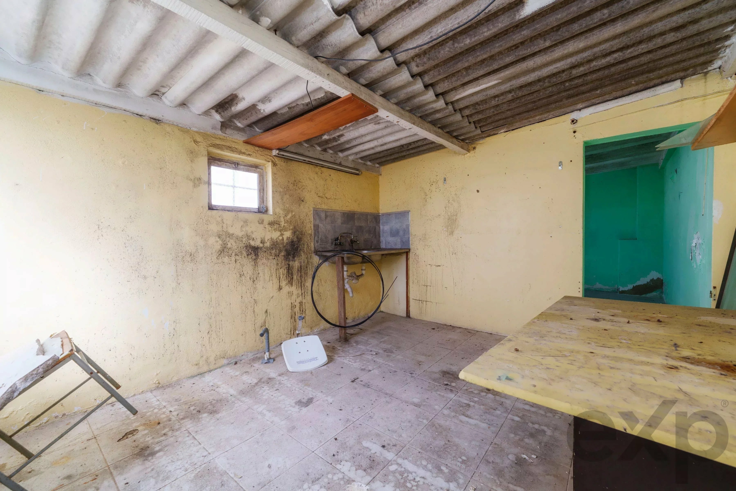 Dusty, neglected interior with yellow walls, grime below, exposed corrugated ceiling, a small window on the left, and scattered debris including a chair and a wooden plank.