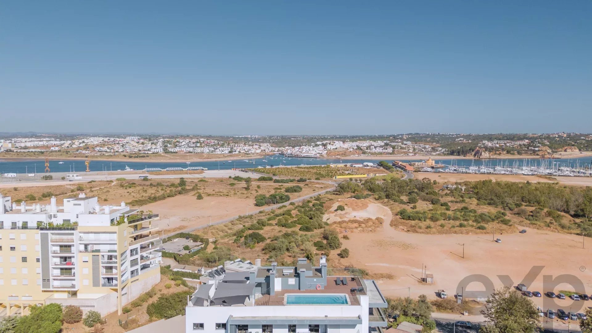 Aerial view of a coastal city with a marina, boats, and white apartment buildings along a sandy, arid landscape under a clear blue sky.