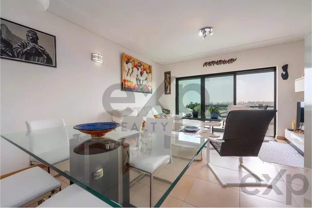 Bright open-plan living and dining room with a glass table, white chairs, and a black recliner by a large balcony window.