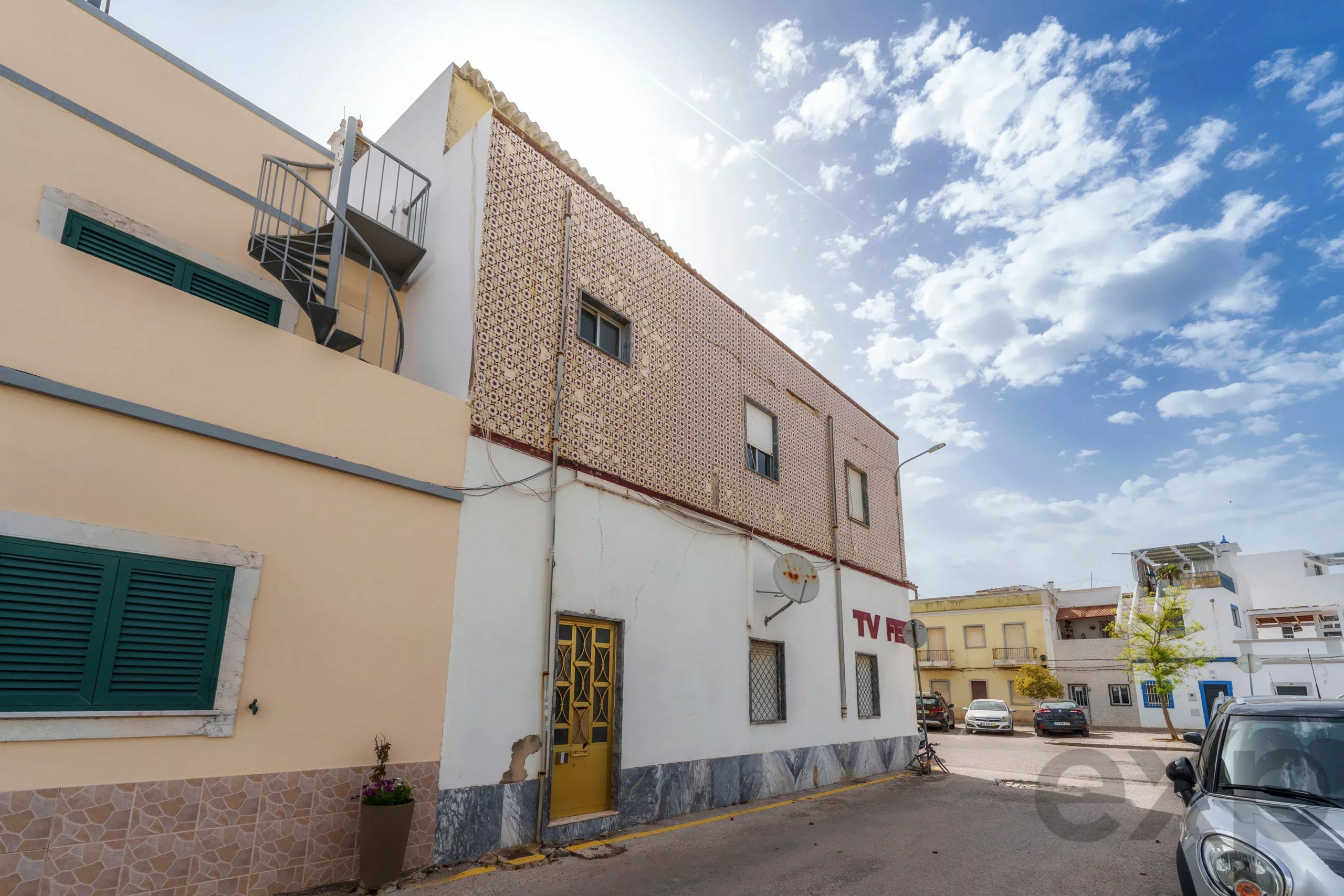 Building with pale yellow walls, green shutters, and a metal spiral staircase against a patterned tiled facade under a blue sky. A street corner showing a beige building with a decorative tiled wall, a yellow door, and parked cars.