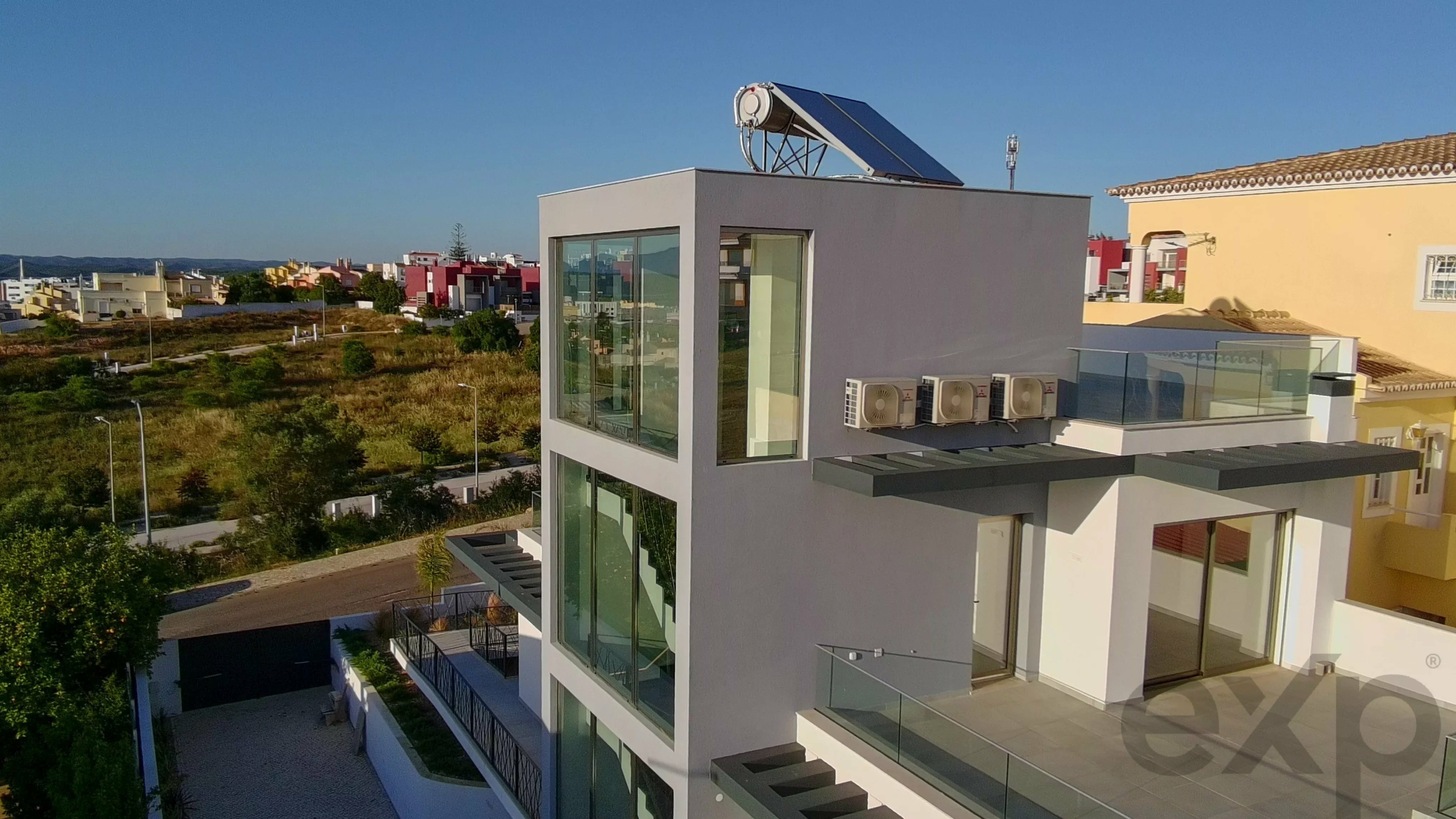 Modern white apartment building with glass balconies, solar water heater on roof, and AC units along the side; suburban neighborhood in background under blue sky