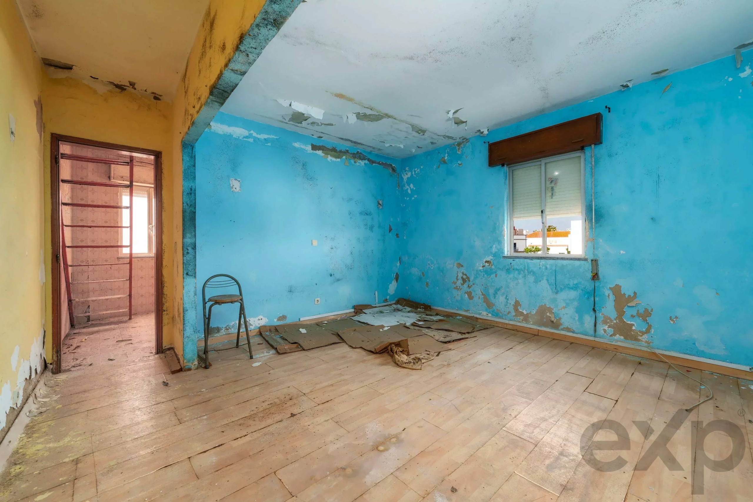 Abandoned room with blue walls and yellow doorway, peeling paint, and debris scattered on a wooden floor, window on the right letting light in.